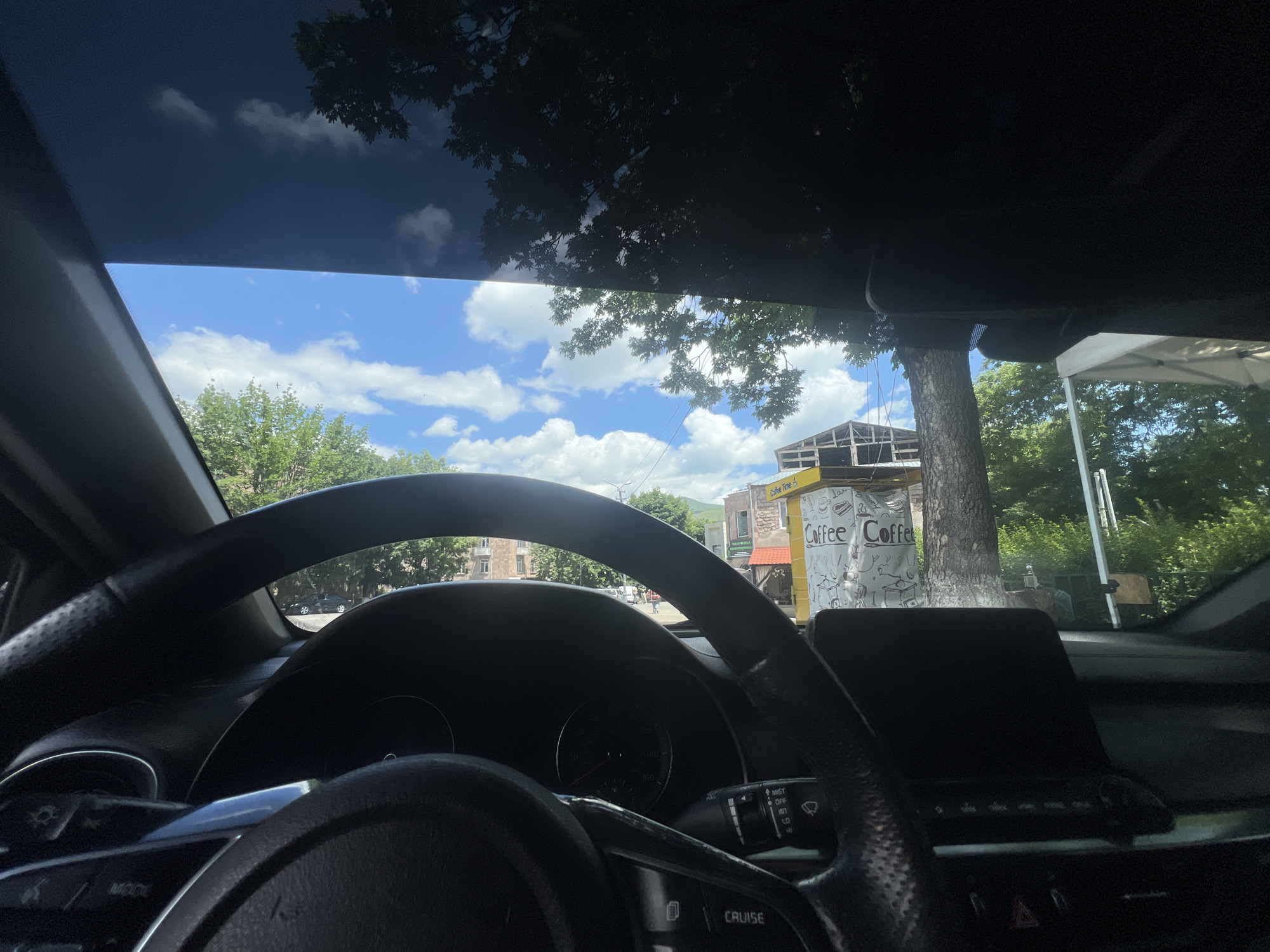 The image is taken from inside a car, looking out through the windshield. A dark tinted panoramic sunroof is visible at the top, partially obscuring the sky. The sky is bright blue with scattered white clouds, and sunlight filters through the leaves of a large tree in the foreground.
In the middle ground, a street scene unfolds. A coffee stand with the text "Coffee Coffee" is prominently displayed on the right. Behind the coffee stand, a large tree trunk is visible. Further back, buildings with reddish-brown roofs and stone facades can be seen, suggesting an urban environment. The road in front of the buildings appears to be paved. Some people are walking on the sidewalk, and a few cars are parked along the street. The overall impression is a sunny day in a town or city.
The foreground of the image is dominated by the interior of the car. The steering wheel is large and black, with control buttons visible. The dashboard and a digital display screen are also present. The driver's side controls for lights and wipers are visible on the left side of the steering column. The image suggests the car is parked or moving slowly on a street in Vanadzor, Armenia, given the architectural style and the presence of a coffee stand commonly found in urban areas.