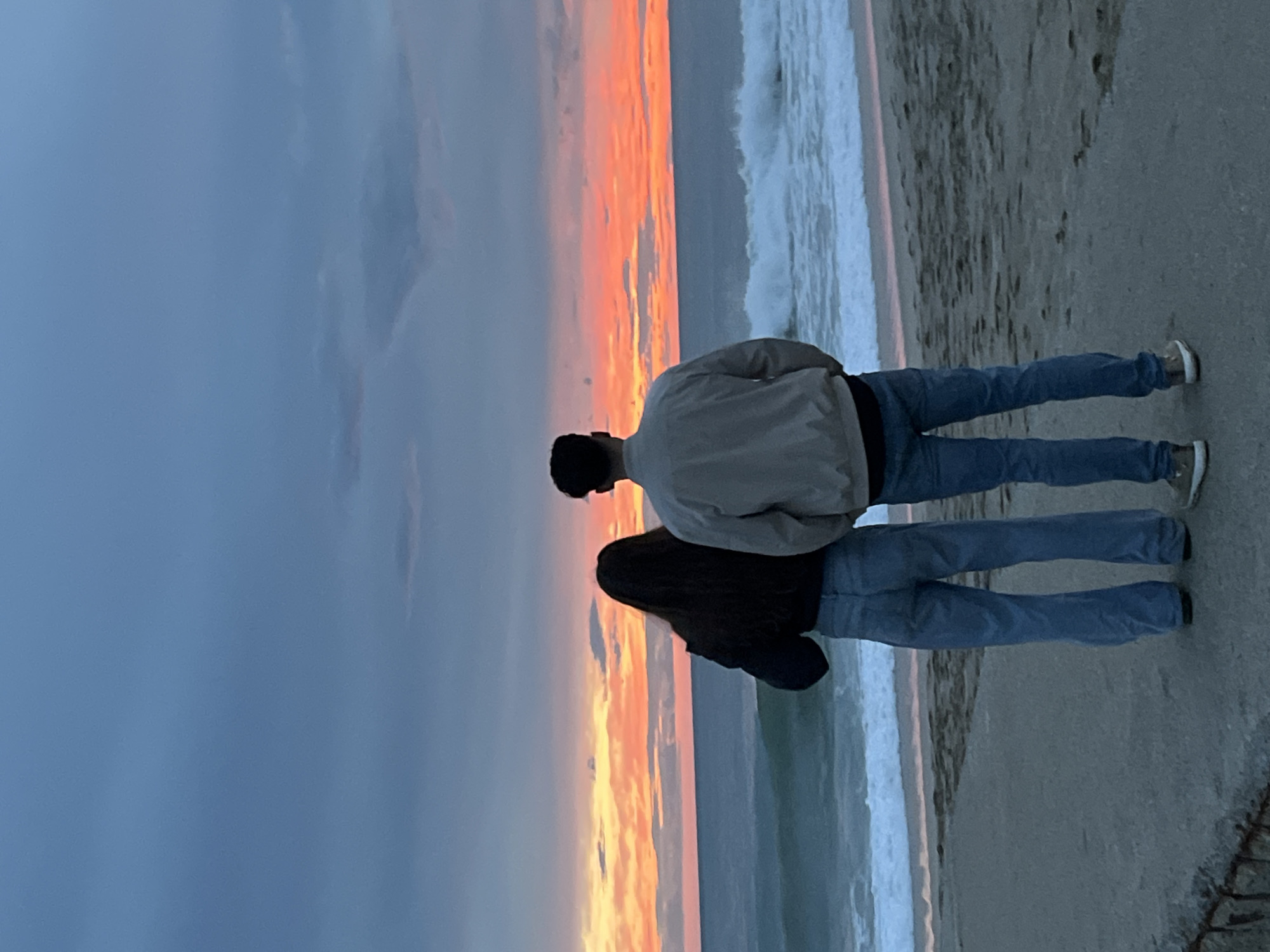 A couple is standing on a beach, looking out at the ocean and a colorful sunset. The man is wearing a light-colored jacket and blue jeans, while the woman is wearing a dark jacket and blue jeans. The waves are crashing on the shore, and the sky is filled with orange, pink, and gray clouds. The location appears to be a beach; it does not allow an identification of a place in Alanya, Turkey.