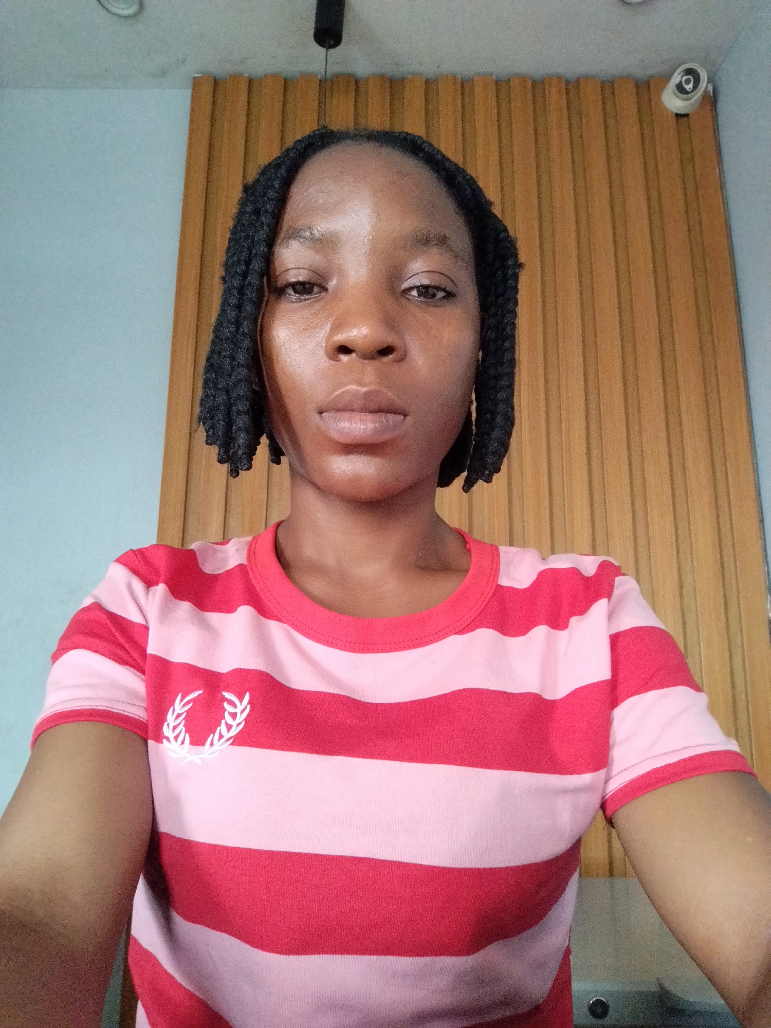 A young Black woman with braided hair and a striped t-shirt is captured in a self-portrait, likely taken indoors in Uselu, Nigeria, given the visual cues. The background features a wall with vertical wooden panels and a light blue wall section. A security camera is visible mounted on the upper right corner of the wooden paneling, and a light fixture hangs from the ceiling in the upper center. The woman is looking directly at the camera, with her face slightly illuminated. Her t-shirt is pink and red striped, with a white laurel wreath logo on the left chest. The overall impression is that of a casual, personal photograph taken in a domestic or semi-public indoor space.