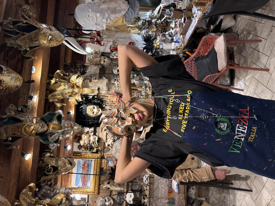 The image depicts the interior of a Venetian mask workshop or store in Venice, Italy. A woman stands centrally, posing with a decorative half-face Venetian mask featuring gold, orange, and purple accents. Her arms are raised with hands clasped behind her head. She wears a black t-shirt emblazoned with yellow text "EVERYTHING YOU LIKED FIVE YEARS AGO," beneath a paint-splattered navy blue apron embroidered with "VENEZIA ITALIA" and a laurel wreath. A silver chain necklace hangs over her attire. Partially visible behind her to the left is a second individual with grey hair, wearing a tan jacket. The background is densely filled with a vast assortment of traditional and ornate Venetian masks, including Jester, Bauta, and Plague Doctor styles, displayed on walls and hanging from dark wooden ceiling beams. A framed painting of a Venetian canal scene is visible on the left wall. Worktables with crafting supplies and more masks are present on both sides, and a salmon-pink chair with cushions is visible on the right. Spotlights illuminate the scene from the ceiling.