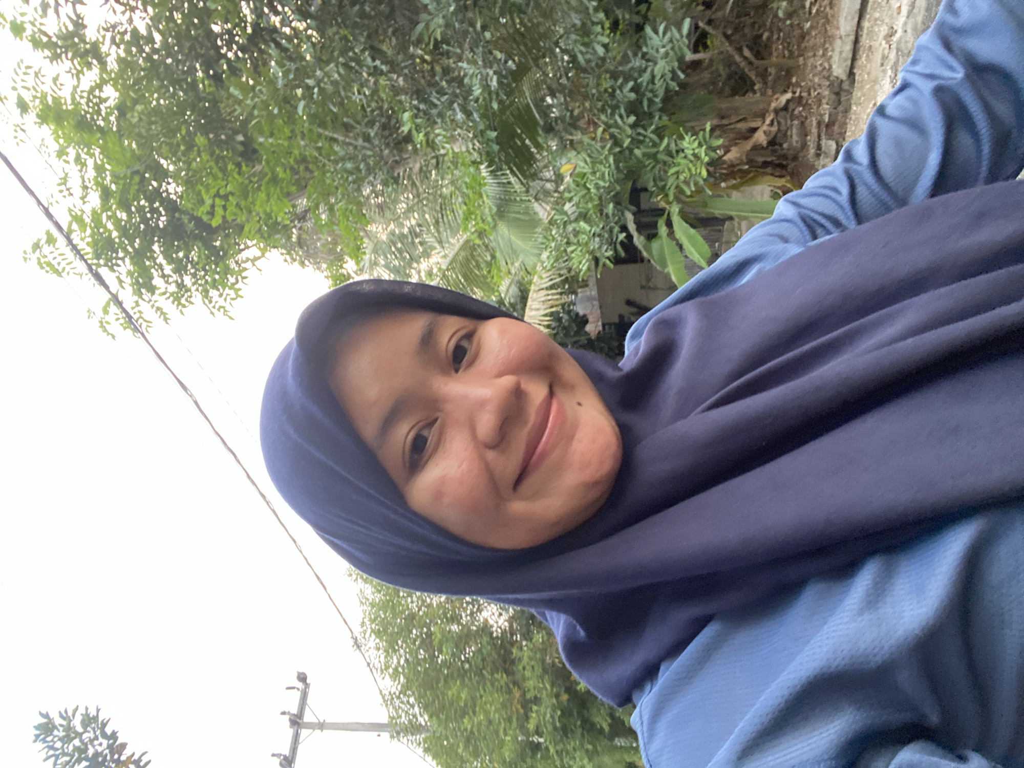 A close-up selfie shows a woman wearing a dark blue hijab and a light blue shirt. She is smiling and looking at the camera. The background is out of focus but appears to be an outdoor environment with lush green trees and foliage, suggesting a location in a tropical or subtropical region, possibly within the city of Banjarbaru, Indonesia, known for its green spaces. A utility pole with wires is visible in the upper left portion of the frame. The lighting suggests it is daytime, possibly late afternoon, with soft, diffused light.