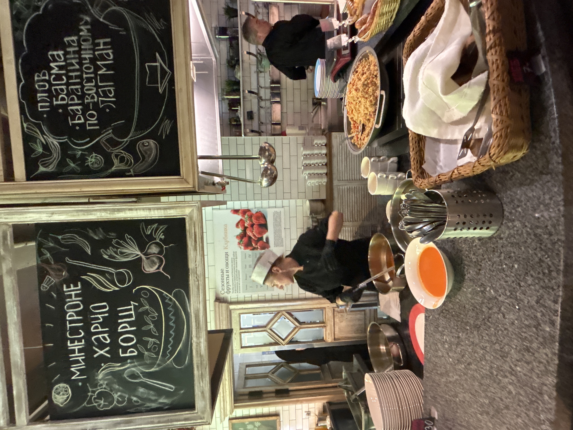 This media file captures a lively scene inside a food establishment, likely a restaurant or a catering service, as suggested by the presence of food preparation areas and serving dishes. The image is oriented vertically, with the main action taking place in the middle ground and background.
In the foreground, two chalkboards display handwritten menus in white and pink chalk. The left chalkboard lists dishes like "Минестроне," "Харчо," and "Борщ," along with drawings of vegetables and a bowl. The upper chalkboard, to the left, lists "Плов," "Басма," "Баранина по-восточному," and "Лагман," accompanied by drawings of ingredients and a steam-venting pot.
The middle ground features a chef wearing a black uniform and a white chef's hat, actively engaged in food preparation. The chef is seen stirring a pot with a ladle, suggesting a cooking or serving process is underway. Behind the chef, a tiled wall with shelves holds various items, including stacked cups and what appears to be a basket of bread. A poster with images of strawberries and text in Cyrillic is also visible on the wall.
The background shows more of the restaurant's interior, with glimpses of kitchen equipment and possibly more service areas. The overall atmosphere appears busy and functional, with the focus on food and its preparation. The lighting suggests an indoor setting, possibly during daylight hours. The visible text, written in Cyrillic, indicates the location might be in a Russian-speaking region. The scene evokes a sense of culinary activity and the presentation of food.