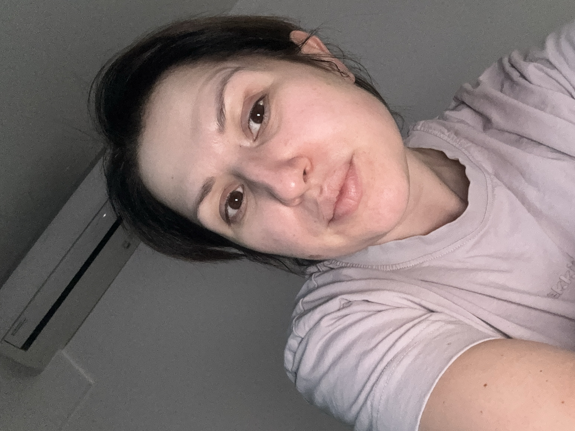 A close-up selfie of a woman with dark hair and brown eyes. She is wearing a light-colored t-shirt. An air conditioning unit is visible in the upper left corner of the frame. The environment is a dimly lit indoor space. The location is unknown and cannot be determined from this image.