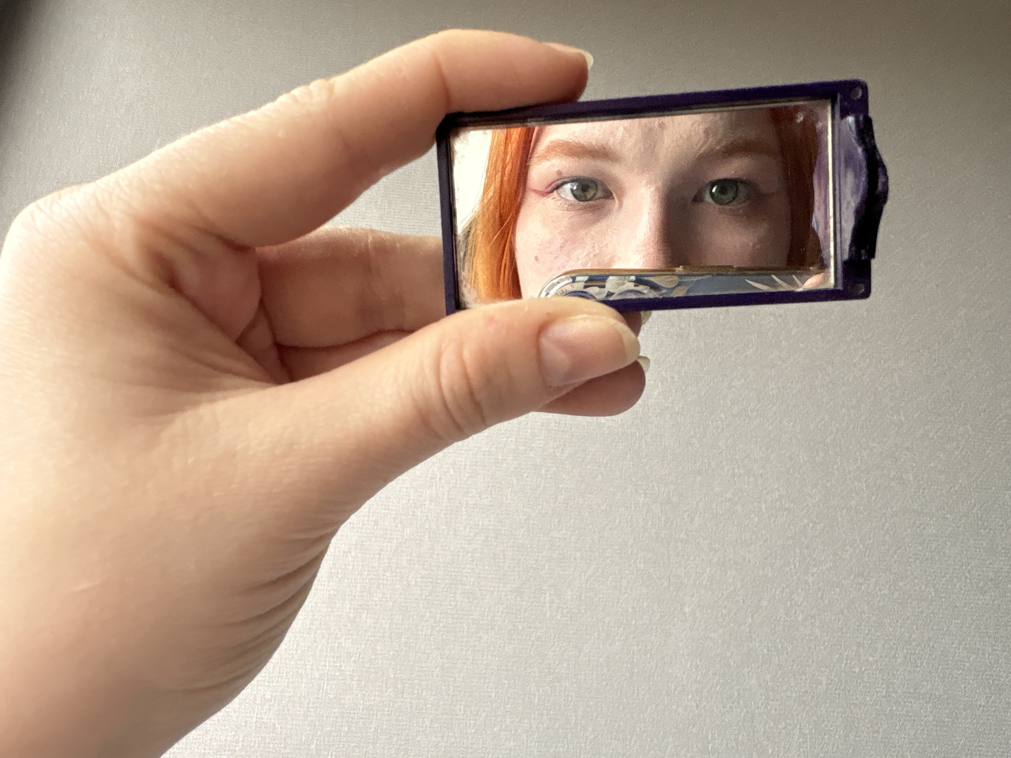A hand holds up a small purple-framed mirror reflecting a person's face. The reflection shows the person has red hair, light skin, and is wearing red eyeliner. The mirror partially obscures their face, covering the lower half of their nose and upper lip. The background is a textured, light grey or white wall. The location is unknown.