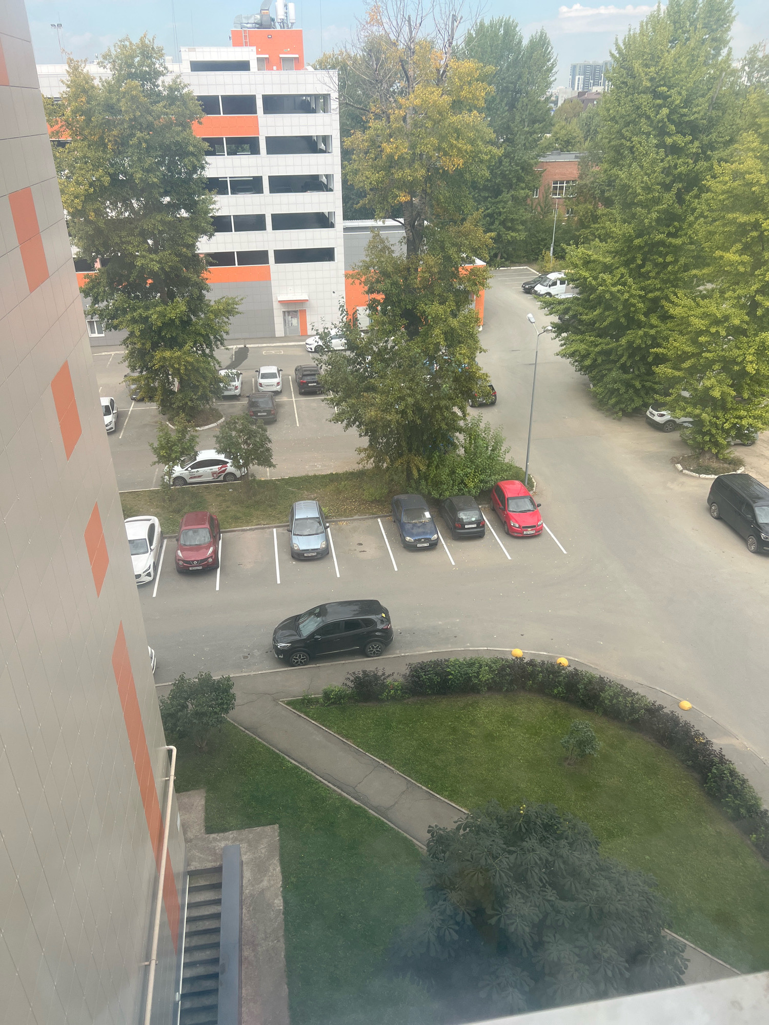 This is a high-angle, full outdoor shot of a parking lot and surrounding area in Kazan, Russia, taken on a cloudy day. A multi-story parking garage with orange and white accents is on the left. The ground level of the garage has parked cars. A paved lot with white parking lines is visible in front of the garage and to the right, with numerous cars parked. Trees with green and yellowing leaves are scattered throughout the scene, some bordering the parking lot. A sidewalk and a grassy area with bushes and trees are also present. A darker, more modern-looking SUV is parked in the foreground right. The buildings are modern and appear to be in a residential or commercial district.