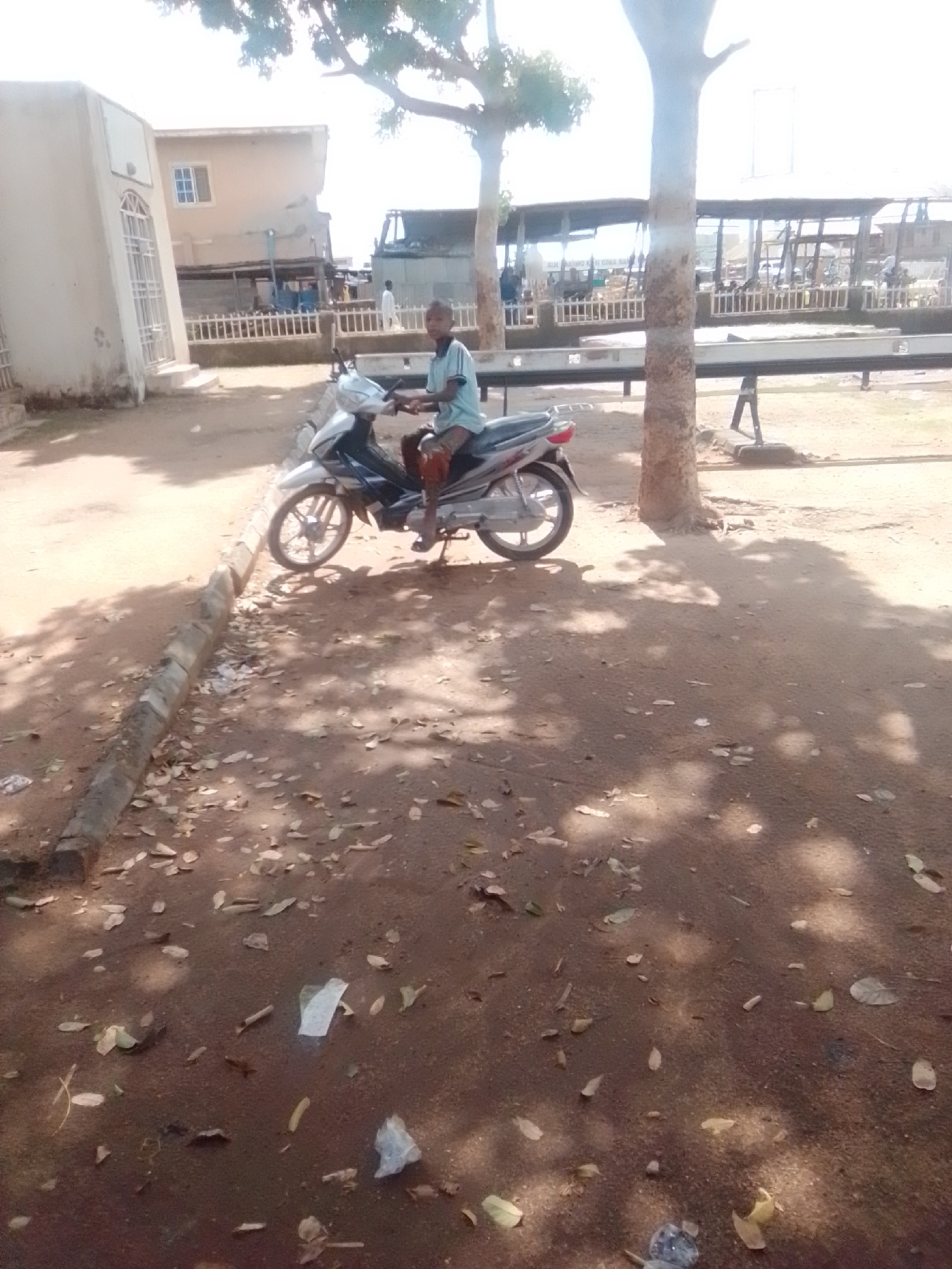 A young boy, wearing a blue t-shirt and shorts, sits astride a silver and blue motorcycle. He is looking forward, his hands on the handlebars. The motorcycle is parked on a dirt road, with a curb to its left. In the background, there are several buildings and a covered market area. The image appears to have been taken on a sunny day, with shadows cast by trees on the ground. The overall setting suggests an outdoor environment in a town or city. The location is the city of Gombe, Nigeria.