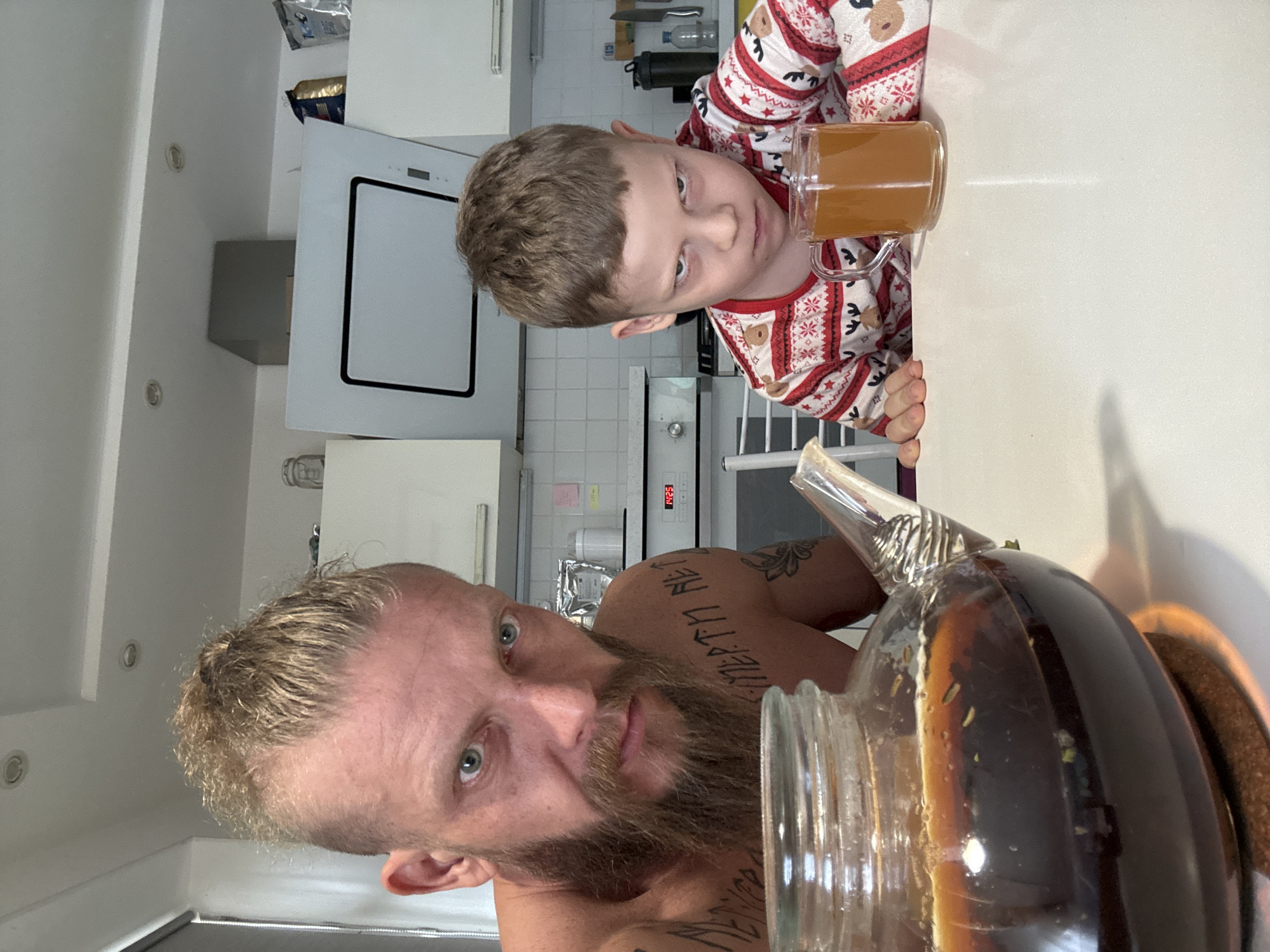 A shirtless man with blond hair, a beard, and tattoos poses with a glass teapot. Next to him, a boy in Christmas pajamas holds a mug of liquid. The background shows a white kitchen setting with appliances. There is no indication of Volzhsky, Russia.