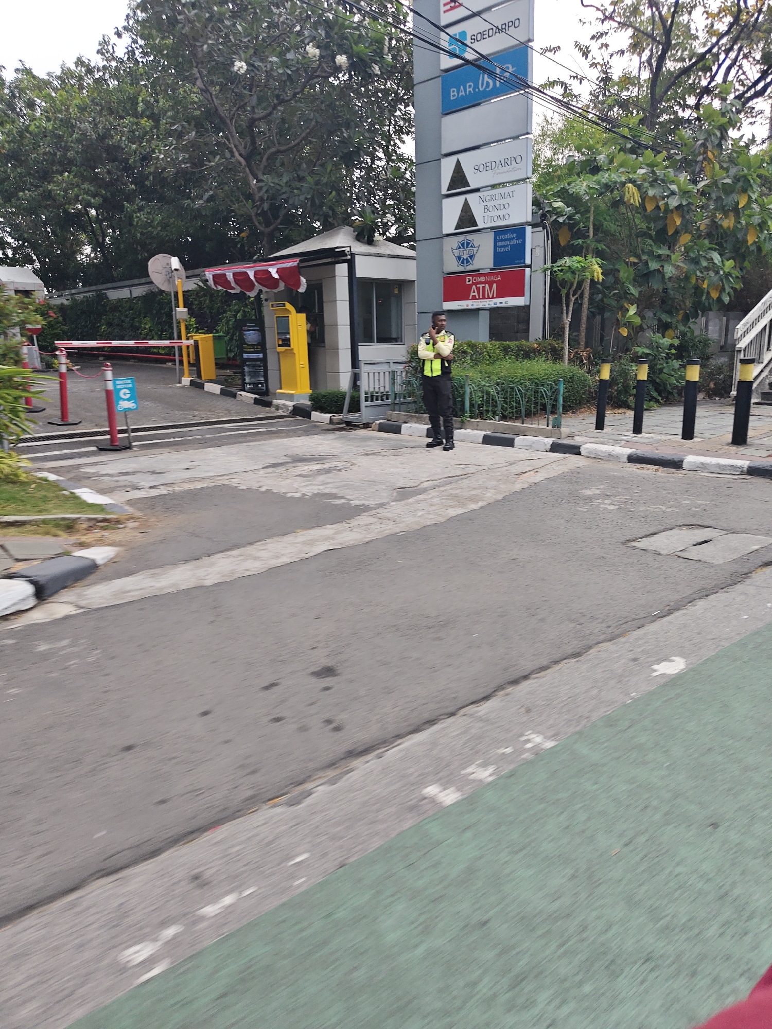 The image depicts an outdoor scene in West Jakarta, Indonesia, featuring a security guard standing near a ticket booth and a signpost with various logos. The security guard, dressed in a high-visibility vest over a dark uniform and pants, is standing on the right side of the road, facing left and holding what appears to be a mobile phone.
To the left of the guard is a small ticket booth with a red and white striped awning. Behind the booth, a yellow ATM machine is visible. Further left, a red and white barrier gate is partially lowered across the road, with bollards and red poles indicating a restricted area. A blue sign with a white motorcycle symbol is also present near the barrier.
A tall signpost stands to the right of the ticket booth, displaying multiple logos and text, including "SOEDARPO," "BAR," "SOEDARPO Foundation," "NGRUMAT BONDO UTOMO," a logo with the text "creative innovative travel," and "CIMB NIAGA ATM."
The environment includes a paved road with distinct white and black painted curbs, suggesting a traffic control or pedestrian area. Greenery, including trees and bushes, surrounds the area, with some trees exhibiting yellowing leaves. The sky is overcast. The perspective suggests the image was taken from a moving vehicle, as indicated by the blurred foreground.