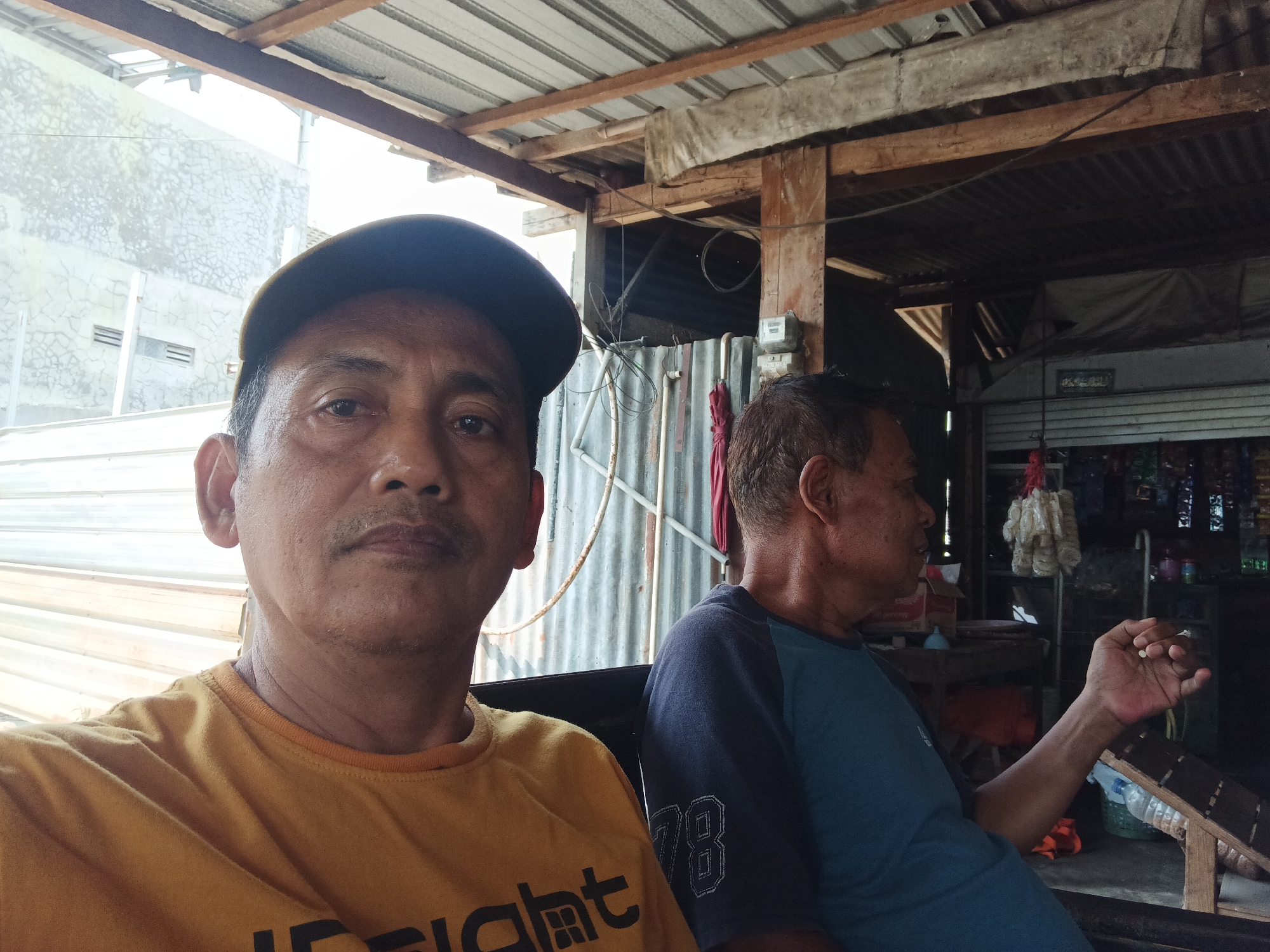 Two middle-aged Indonesian men are seated in what appears to be an outdoor seating area of a small shop or home in Batursari, Indonesia. The man in the foreground, wearing a yellow t-shirt and a dark baseball cap, is looking directly at the camera. He has a mustache and is positioned to the left side of the frame. To his right, another man, wearing a blue and gray t-shirt with "78" visible on the sleeve, is looking away from the camera, with his right hand raised and holding what appears to be a small, light-colored object. Behind the men, the environment suggests a modest, possibly rural or semi-urban setting. A corrugated metal wall is visible behind them, along with wooden beams and a corrugated metal roof. To the right, shelves stocked with various items, possibly snacks or household goods, are visible within the structure. The overall impression is one of a casual, everyday scene.