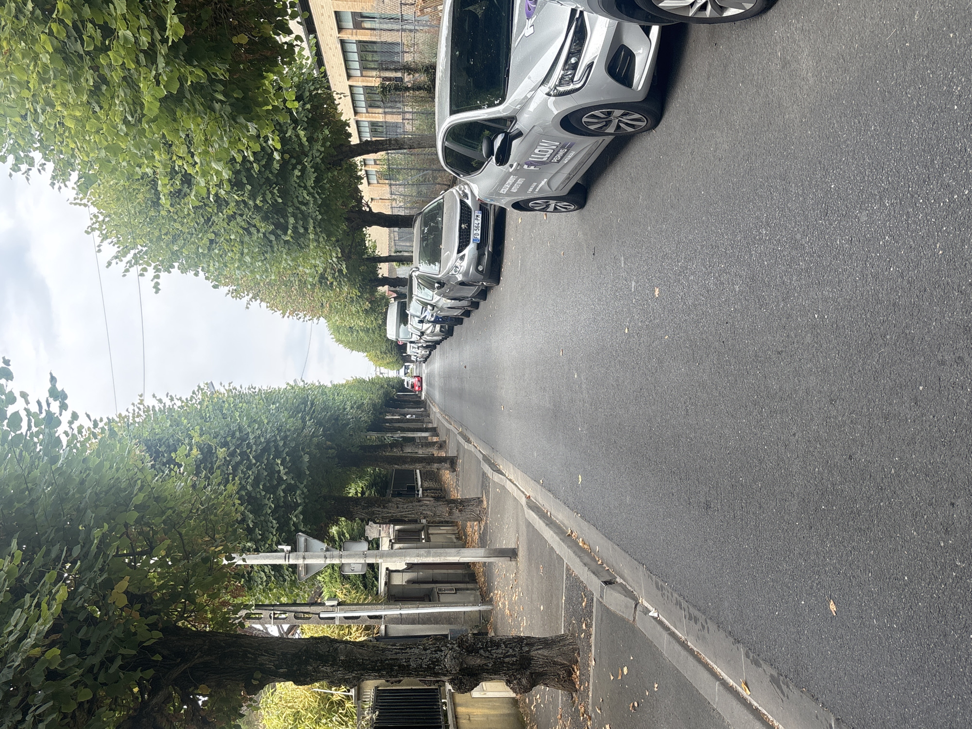 The image depicts a residential street in Aulnay-sous-Bois, France, lined with trees and parked cars. The road is paved and appears to be a two-way street. On the left side of the street, there is a sidewalk with a curb. Several mature trees with dense green foliage are planted along the sidewalk, casting shadows on the pavement. Some fallen leaves are scattered on the sidewalk and the street. The buildings visible on the right side of the street are brick structures with multiple windows.
Several cars are parked in a line along the right side of the street. The closest car, a silver Renault Clio, has "FOLLOW ECOLE DE CONDUITE AUTO ECOLE" and "PERMIS AUTO ECOLE" branding on its side. Further down the street, other cars are parked, including a silver Peugeot with a license plate reading "FD-564-PM". A red vehicle is visible in the distance, at the end of the street.
The sky is overcast, suggesting a cloudy day. There are no people visible in the image. The overall scene is that of a quiet, tree-lined urban street with vehicles parked along the side.