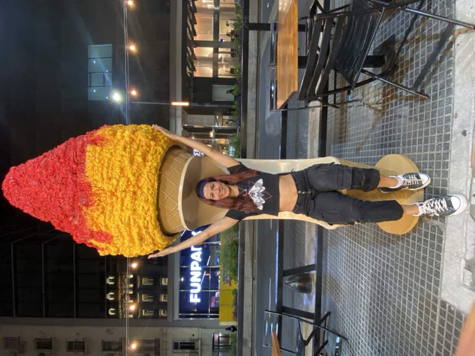 A woman lies inside a giant ice cream cone sculpture on a city street at night. The cone is red and yellow, with a waffle cone pattern. The woman wears a black crop top, dark pants, and sneakers. She is smiling and has her arms outstretched to the sides of the cone. In the background are buildings, streetlights, and outdoor seating for a cafe. The word "FUNPAR" is visible on a building sign.