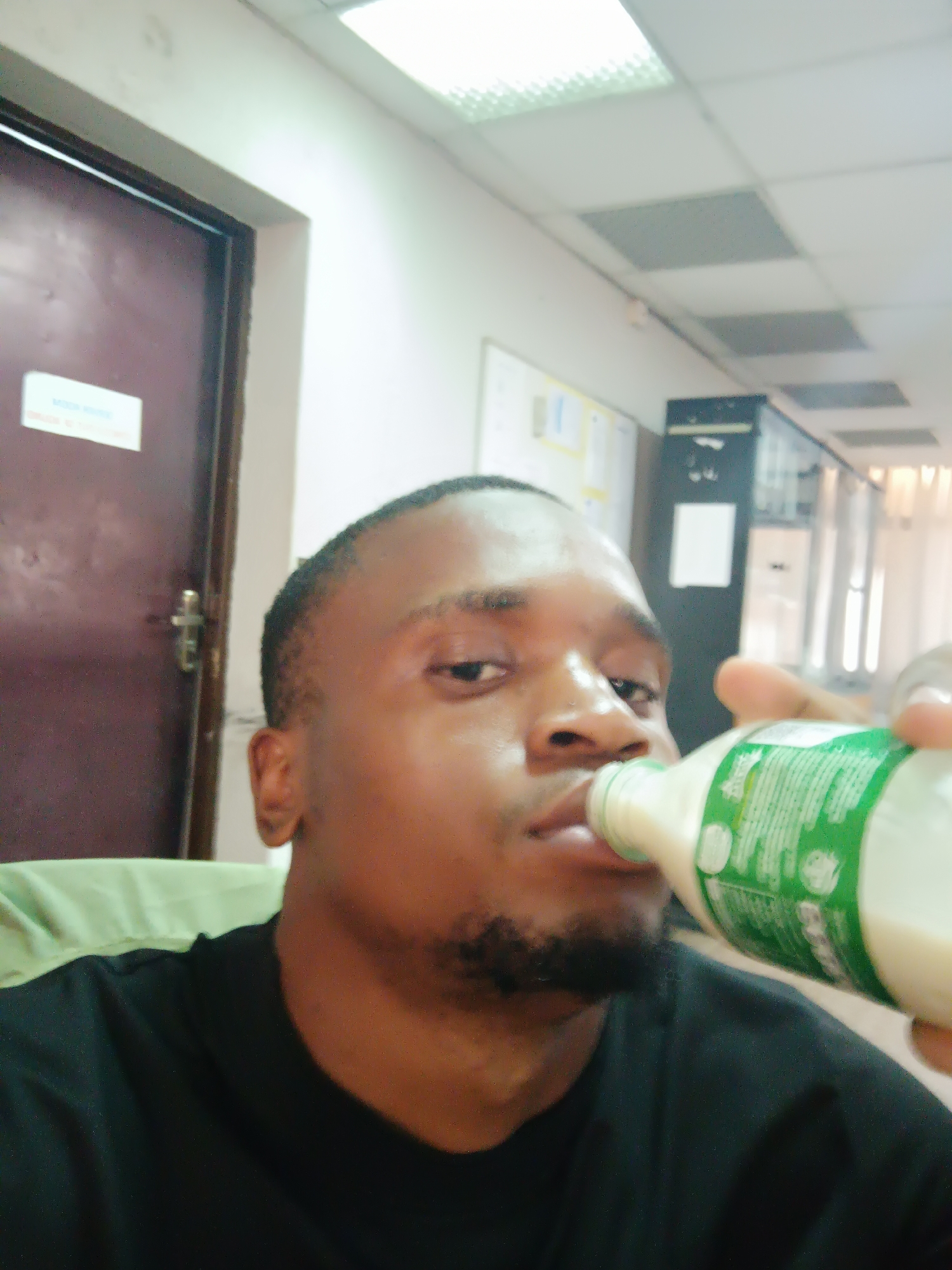 An indoor, eye-level shot features a man drinking from a bottle, positioned slightly to the right of the frame. He has short dark hair and a dark beard or stubble, and is wearing a black t-shirt with a light green collar or shoulder section. His gaze is directed towards the camera with a neutral expression as he brings a white bottle with a green label to his lips. The bottle contains a white liquid, likely milk or a dairy drink, and its label has small, unreadable text and green symbols.
The setting appears to be an office or institutional environment. To the left, a dark brown door with a silver handle/lock is visible. A rectangular white sign on the door displays "MODE OF ENTRY" and smaller, unreadable text below. The background includes light-colored walls, with acoustic ceiling tiles (some white, some gray) and fluorescent light fixtures visible above. Behind the man's right shoulder, a bulletin board or white board with papers is partially visible, along with a dark piece of office furniture or equipment further back. A window or glass partition with vertical elements is dimly discernible in the far right background. The image is taken in Kakuri Gwari, Nigeria.
