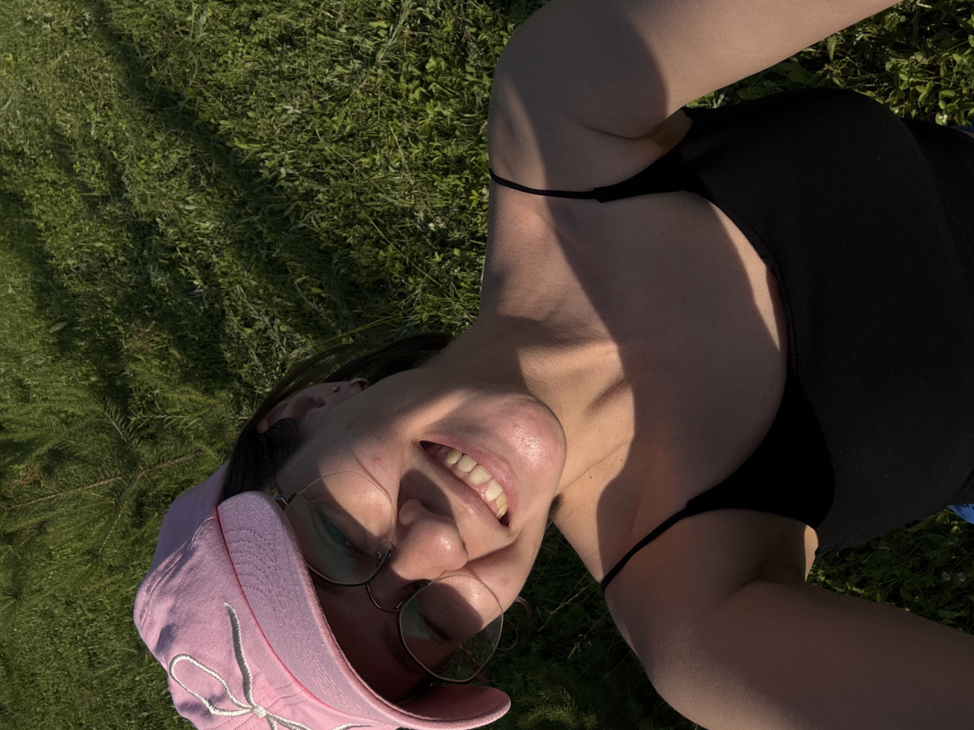 A young woman wearing a pink baseball cap, eyeglasses, and a black tank top smiles at the camera while lying on a grassy area. The lighting suggests it is daytime, possibly late afternoon due to the shadows. The background is filled with green grass and some tall, thin plants. There is no discernible indication of the woman's location within the city of Doksha, Russia.