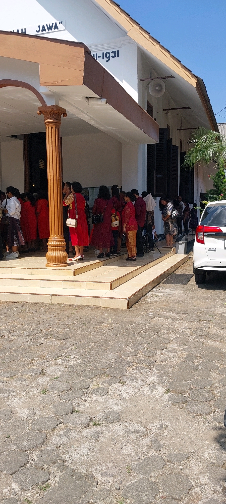 The image captures a sunny day scene outside a building with a prominent brown-trimmed awning supported by a fluted, golden-brown pillar. The building's exterior displays the inscription "ASIA JAWA" on a white banner and "II-1931" in black lettering. A group of people, mostly women dressed in traditional Indonesian attire, are gathered on the tiled steps and a ramp leading up to the building's entrance. Several individuals are in shades of red and maroon, while others wear patterned fabrics. A white car with the "ASTRA" logo is parked to the right of the frame. The ground in front of the building is paved with uneven, grey interlocking stones, with sparse vegetation growing through the cracks. The environment suggests a public or institutional setting in Keling, Indonesia, possibly for a cultural event or gathering.