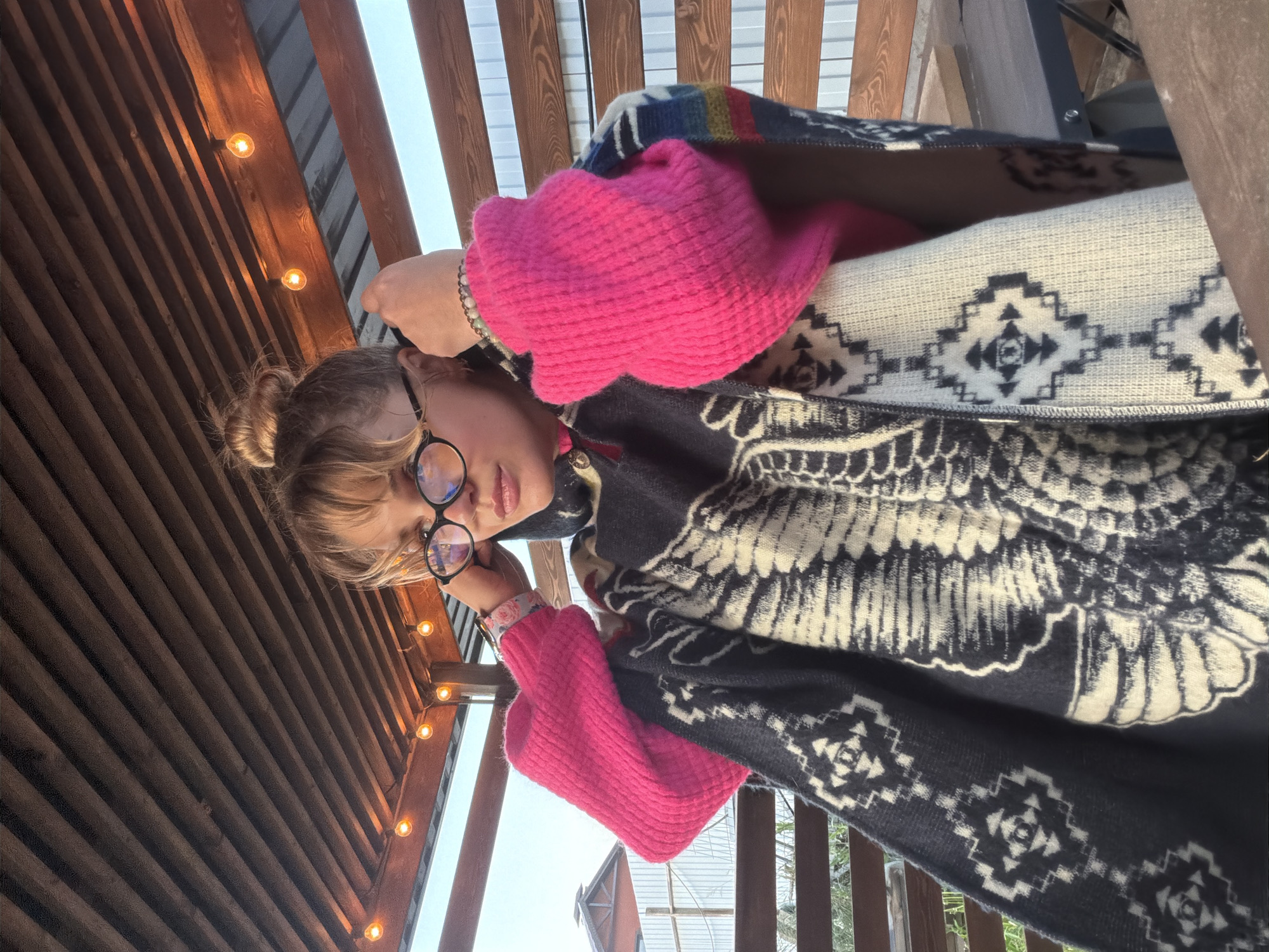 The media file captures a portrait of a young person in an outdoor or semi-outdoor setting. The subject, appearing to be a female, has blonde hair styled in a messy bun and wears black-framed glasses. She is adorned with a pearl bracelet on her left wrist and is dressed in bright pink knit sleeves or a sweater, covered by a thick, dark blue or black blanket or poncho featuring a prominent white eagle-like design on the upper part and geometric patterns composed of diamonds and arrow shapes along the lower edge. Her hands are raised, resting near her face and glasses, and she looks directly at the camera with a neutral expression.
The background reveals a warm and inviting atmosphere. Above, a wooden slatted ceiling or awning is visible, strung with several glowing string lights, casting a soft, warm illumination. Beyond this structure, a bright, clear sky is visible, suggesting either late afternoon or early evening, or perhaps a bright overcast day where the lights are turned on for ambiance. In the lower right, part of a wooden railing can be seen, hinting at a balcony, porch, or deck setting. Sparse foliage or trees are subtly visible in the distance, completing the serene outdoor scene. No visible text is present in the image. The location is contextualized as Большая Шильна, Russia.