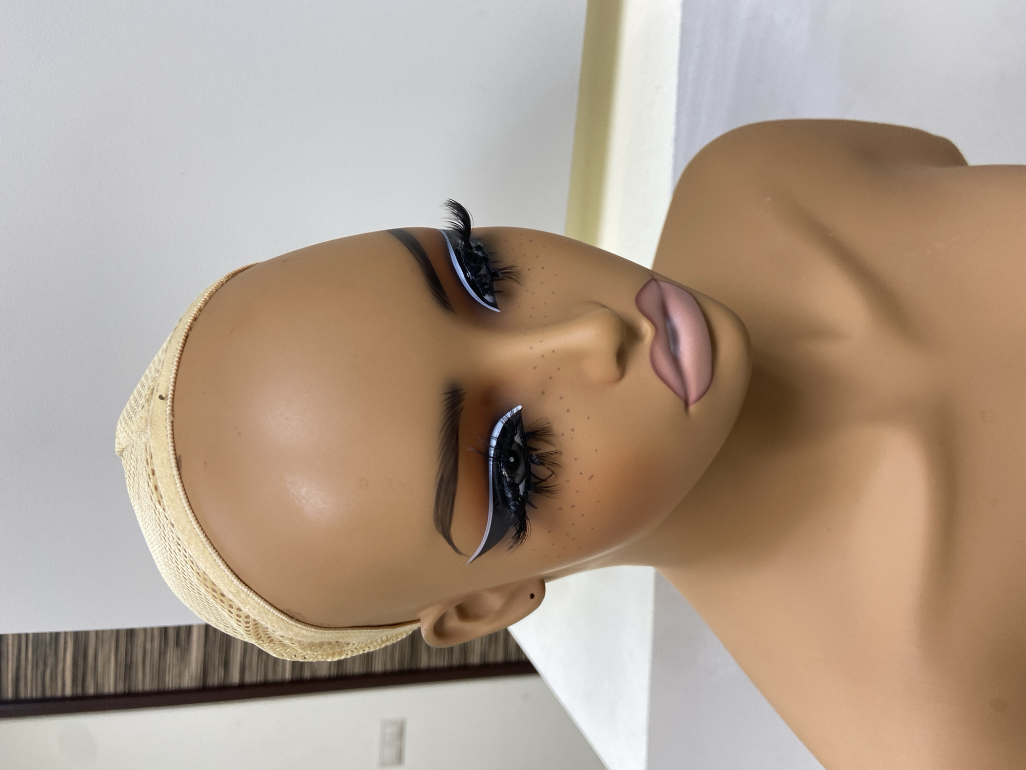 The image shows a tan-skinned mannequin head with elaborate makeup. The mannequin wears a wig cap. Its eyes are heavily styled with long eyelashes and white eyeliner. The lips are painted with a pink and brown gradient. The mannequin appears to be indoors, next to a white wall. There is no information in the image to suggest the location is Kazan, Russia.