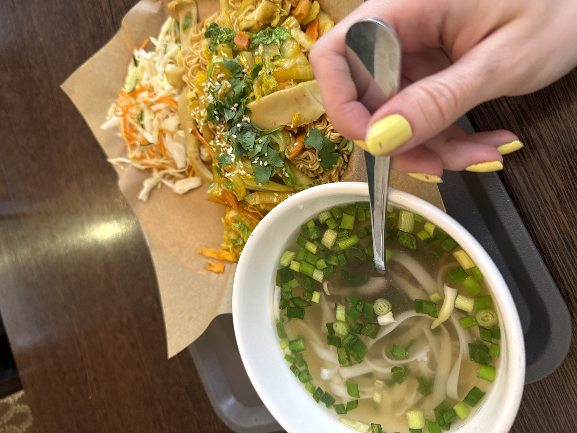 A high-angle shot shows a tray on a dark brown wooden surface, possibly a table. On the tray is a white bowl of soup with green onions, noodles, and a metallic spoon being held by a hand with yellow painted nails. Adjacent to the bowl is a plate with noodles, vegetables, and what appears to be a cabbage slaw. The scene appears to be indoors, possibly in a restaurant. There is no evidence to suggest this scene is in Moscow, Russia.