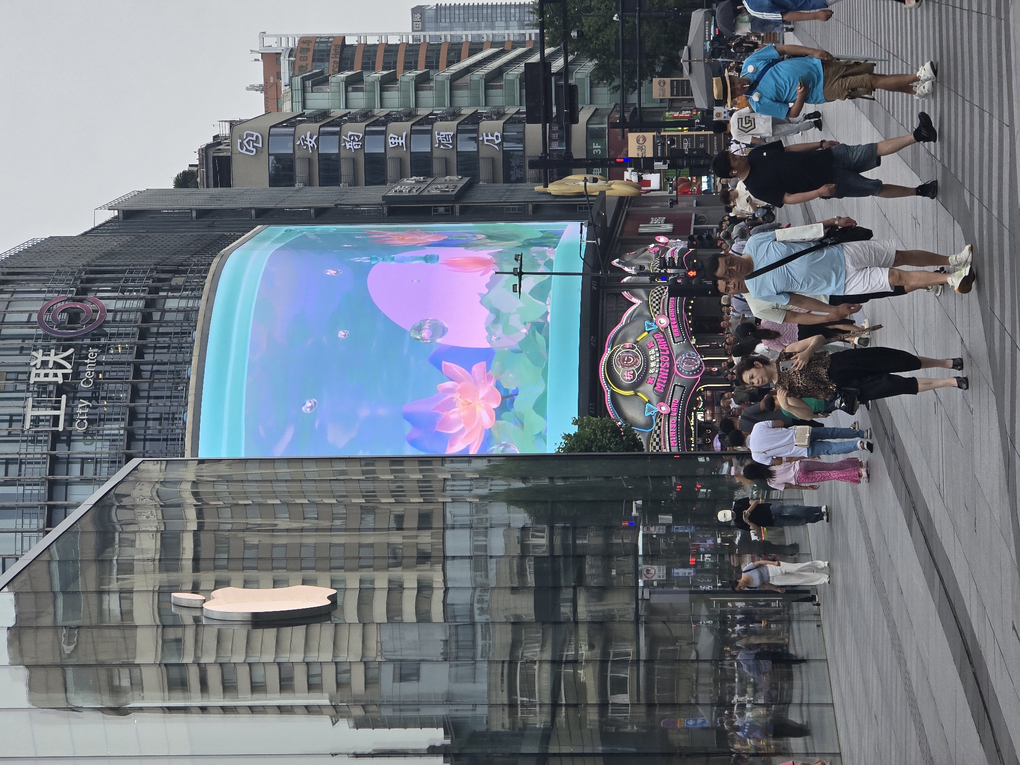 This image captures a vibrant urban street scene, likely a commercial district in Shangcheng District, China. Numerous pedestrians, comprising men and women of various ages, populate the wide paved walkway, some walking purposefully while others appear to be casually strolling or observing their surroundings.
The most prominent feature is a massive, curved outdoor LED screen displaying a vivid, animated scene resembling an aquatic fantasy or an enchanted pond. This display features large pink lotus flowers, bubbles, and a glowing pink orb or moon against a light blue background. To the left, a modern building with a distinctive glass facade prominently displays a large, reflective Apple logo. The building's glass also reflects the surrounding urban landscape and the sky.
Across the street from the Apple store, a multi-story building features a large "MINISOLAND" sign, identifying a Miniso store, along with its Chinese name "名创优品". Other buildings in the background exhibit a mix of architectural styles, some with more traditional-looking roofs and intricate details. Visible text on these buildings includes "工联" (Gōnglián), "City Center", "安韵里酒店" (An Yun Li Hotel), "租" (Rent), "出" (For rent/Exit), "3F", and "国际" (International).
The time of day appears to be overcast daytime, with diffused light and no harsh shadows, suggesting a cloudy or heavily overcast sky. The overall atmosphere is bustling, typical of a busy city center, with people engaged in their daily activities amidst modern commercial displays and architecture.