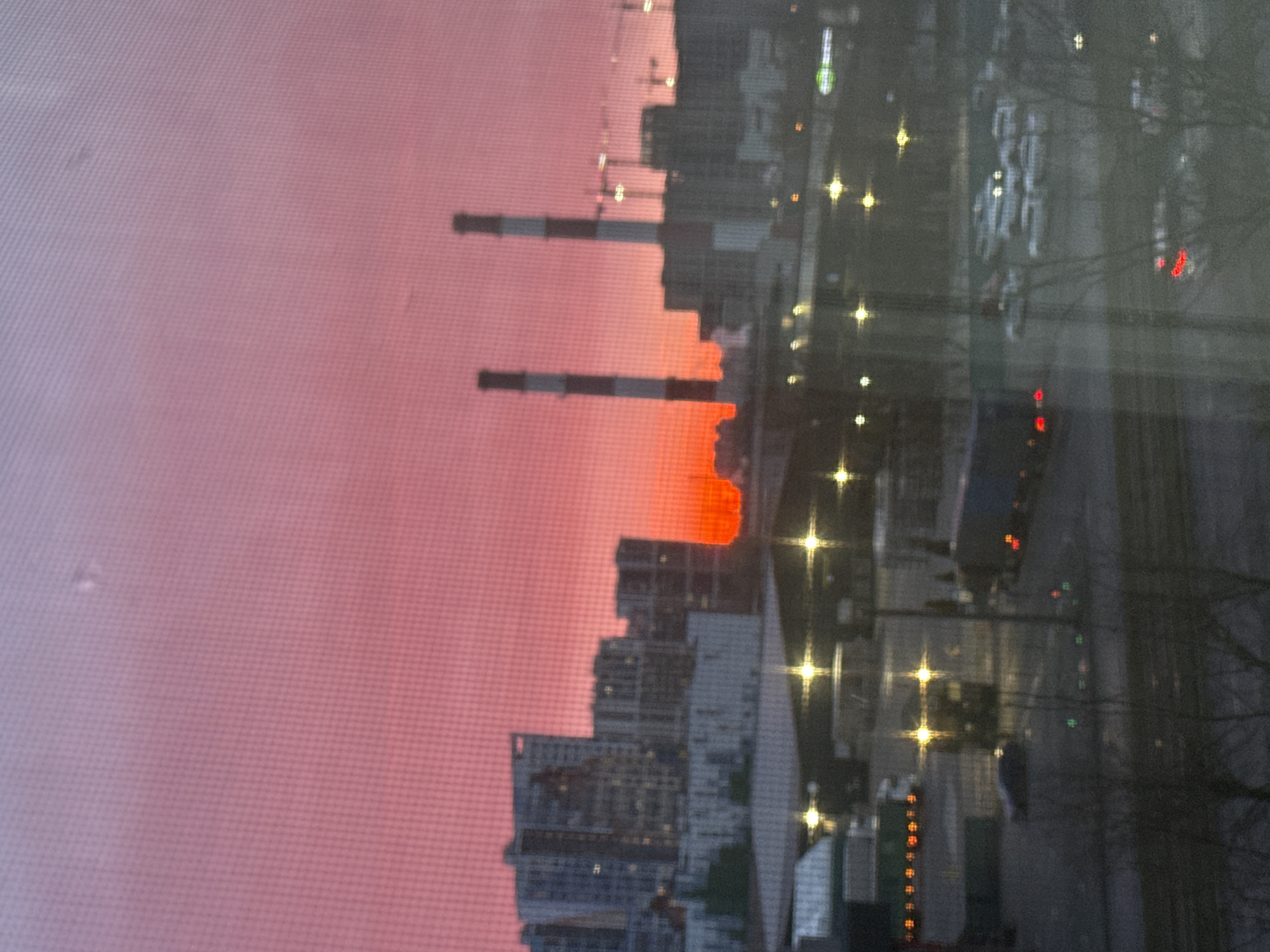 The image depicts a cityscape at dusk or dawn, likely taken through a window due to the slightly blurred effect and the presence of a screen pattern. The sky is a gradient of pink and orange, suggesting a sunrise or sunset. Two industrial chimneys are visible, emitting no smoke.
Below the horizon line, a cluster of high-rise buildings forms a modern skyline. Further down and to the right, a street scene is visible, populated with blurred lights suggesting street lamps and vehicle lights. The image appears to show a view from the city of Saint Petersburg, Russia.