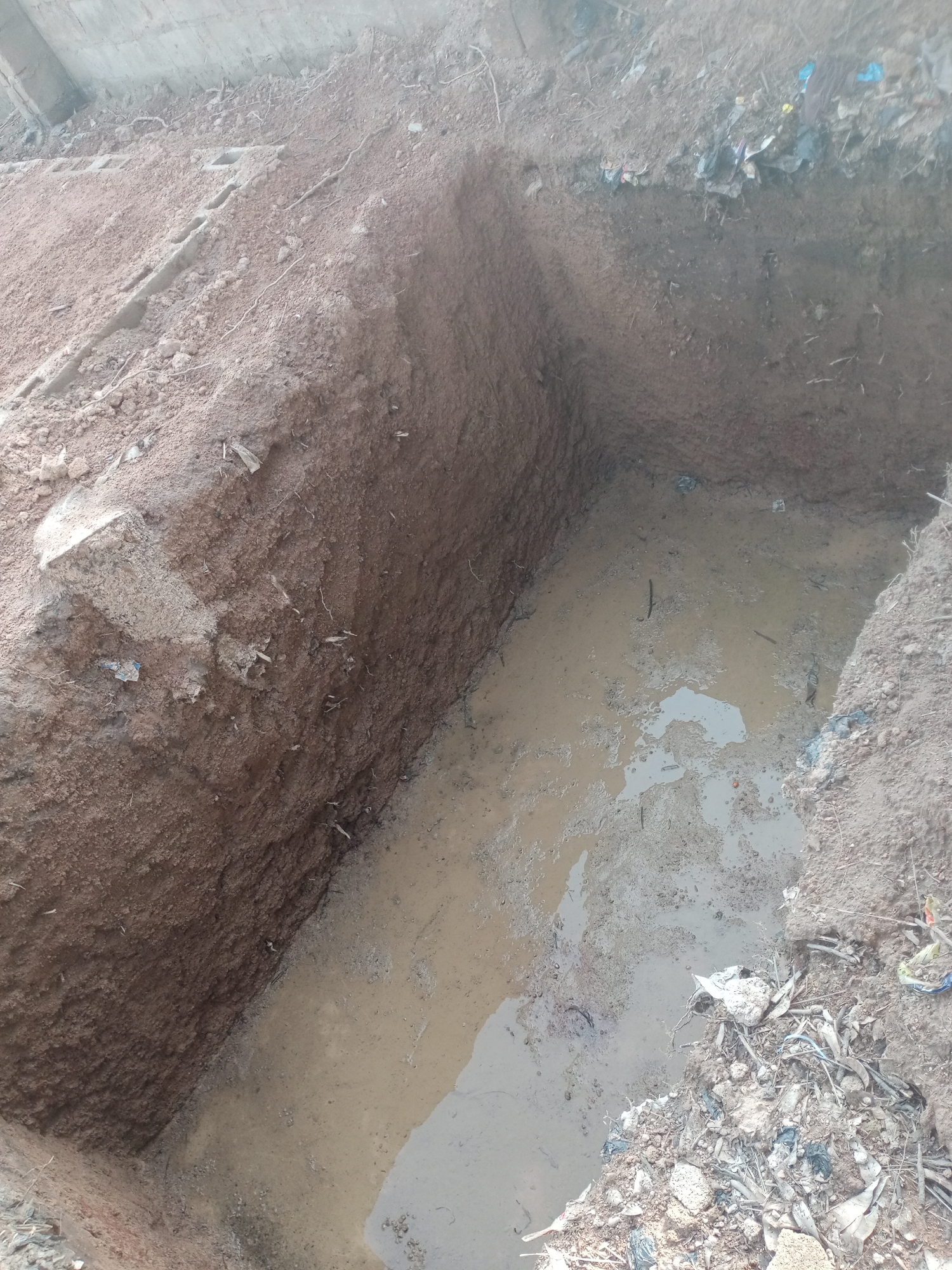 The image depicts an excavated pit filled with murky water and mud. The sides of the pit are composed of reddish-brown soil with visible roots and debris. In the upper left corner, a concrete structure or wall is partially visible, casting a shadow over the pit. The surrounding area appears to be an outdoor, unpaved location, likely in an urban or semi-urban setting, possibly Jalingo, Nigeria, given the context. There are scattered pieces of trash and debris along the edges of the pit, including plastic fragments and dry leaves. The water in the pit is stagnant and appears to be a mixture of rainwater and runoff. No individuals or specific actions are depicted in the image. The overall scene suggests an ongoing construction or excavation project, or potentially a natural accumulation of water in a dug-out area.