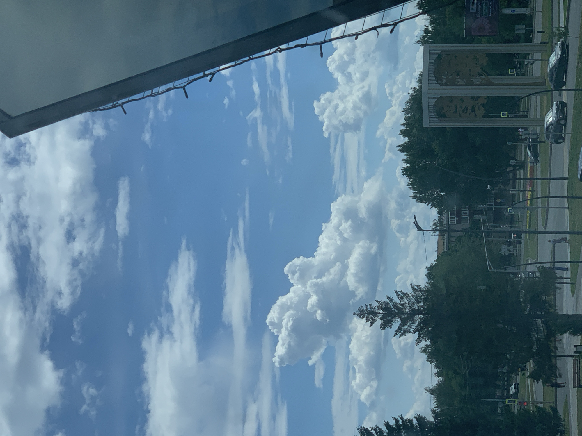 The image depicts an outdoor urban scene under a blue sky with scattered white cumulus clouds. In the foreground, a large, dark green evergreen tree dominates the center. To the right, a portion of a dark, reflective structure with a string of lights along its edge is visible, extending from the top edge of the frame downwards. Behind the tree, a street with several cars is visible. A sculpture featuring two large, abstract tree-like figures within a rectangular frame stands on the right side of the street. Trees and buildings line the background. Several people are seen walking on the sidewalk. The scene appears to be located in Nizhnekamsk, Russia, possibly in a public square or park area, given the presence of the sculpture and trees. The lighting suggests it is daytime, likely late morning or afternoon.