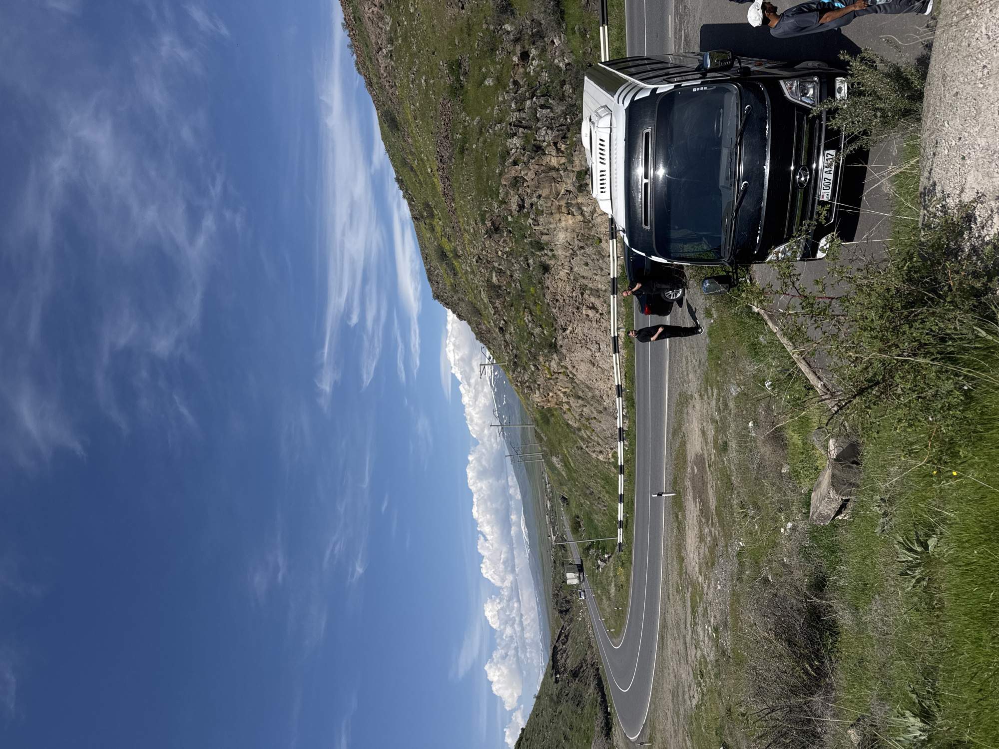 The image shows a black and white van parked on a winding road in Bagravan, Armenia. The license plate of the van reads "007 AT 17". Two people stand near the van. The road is situated along a mountainside covered with greenery. The sky is blue with scattered white clouds. A section of a concrete barrier is visible on the right side of the road. Utility poles run along the distant hills.