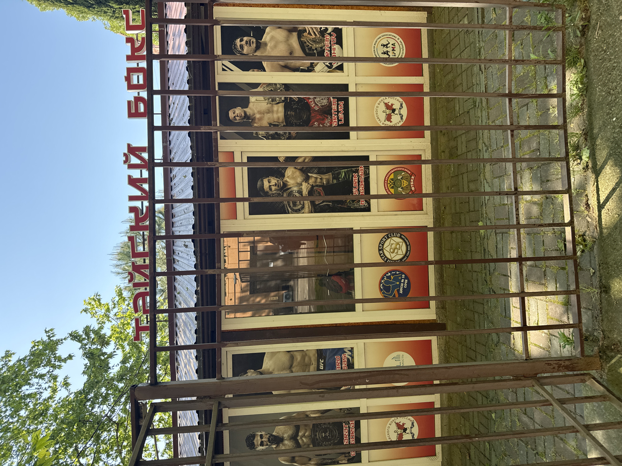 The image shows the exterior of a building, possibly a boxing or martial arts training center in Sochi, Russia. The façade is decorated with posters of fighters and various sport club logos. The text "ТАЙСКИЙ БОК" is visible above the entrance, likely indicating that it offers training in Thai boxing. A metal fence partially obscures the view of the building. The logos on the posters and the text around suggest a focus on martial arts such as Muay Thai, and organizations affiliated with it. The presence of the text "ДЮСШ № 10 СОЧИ" implies this is a youth sports school.
