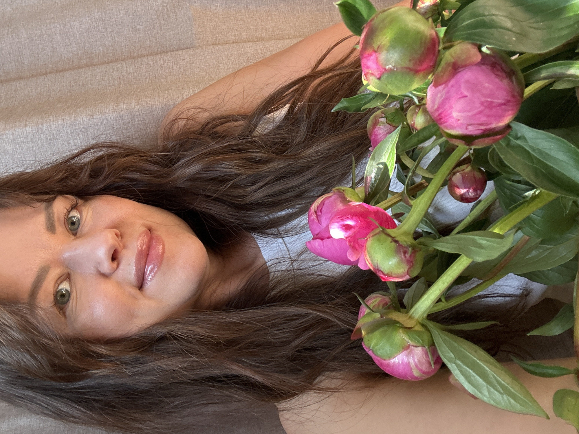 The image shows a close-up portrait of a woman with long, dark, wavy hair, holding a bouquet of pink peonies. She is wearing a white top and her makeup includes lip gloss and eye shadow. The background consists of a textured, beige curtain. The image does not provide any information about the city of Chaikovsky, Russia, or any specific location within it.