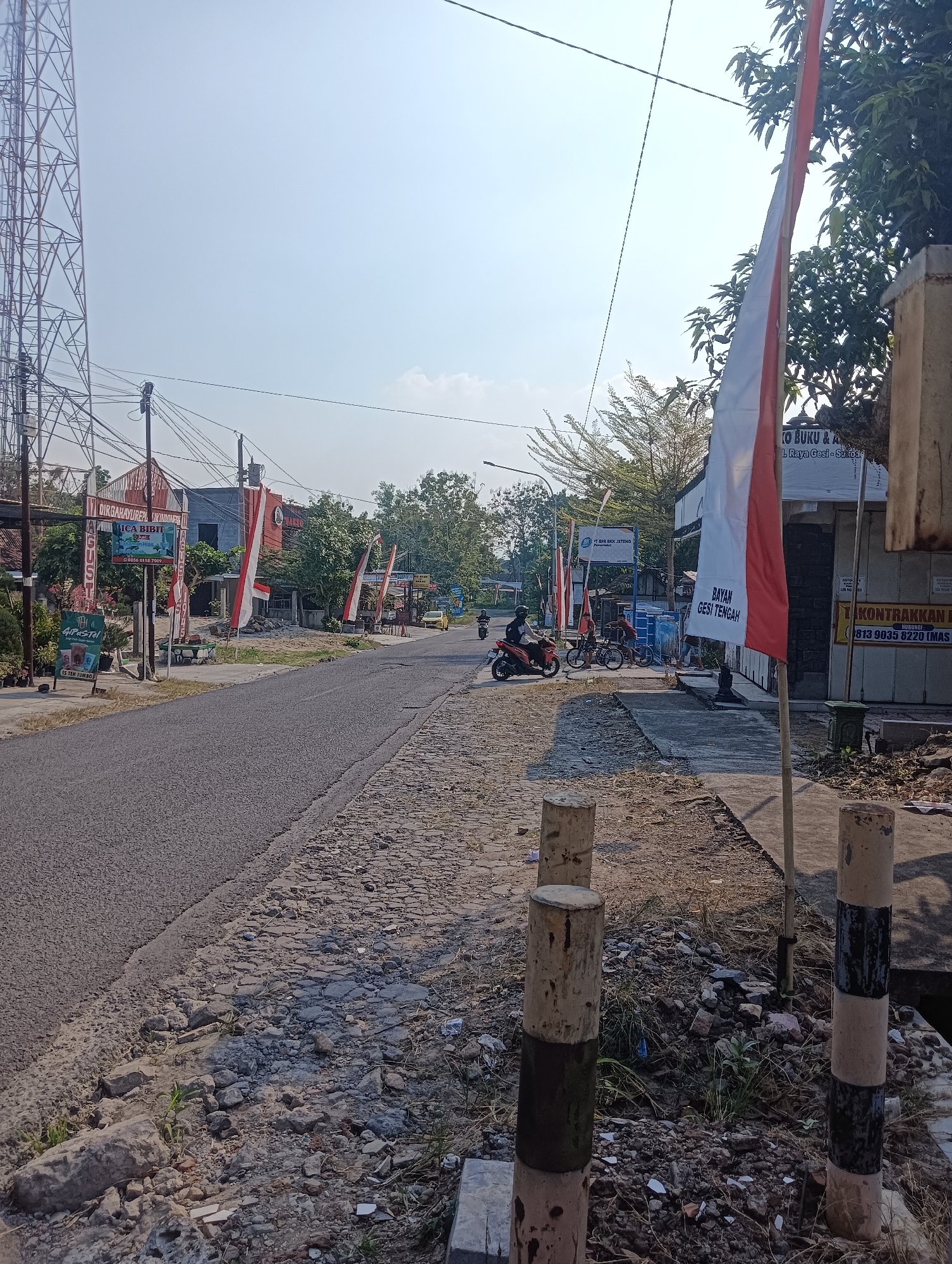 The image depicts a street scene in Palemrejo, Indonesia. A paved road occupies the left and center of the frame, with a rougher, cobblestone-like surface on the right. Several red and white Indonesian flags are prominently displayed along the roadside.
On the left side of the road, a tall communication tower is visible, along with numerous power lines and poles. Several buildings with signs are present, including one with the text "DIRGAHAYU REPUBLIK INDONESIA" and another advertising "4 Pastel". Signs indicating businesses such as "TOKO BUKU & ALAT TULIS" and "KONTRAKKAN" are also visible on the right side of the street.
In the mid-ground, a few individuals are present. One person is riding a motorcycle, and two others are standing near some blue portable water tanks. A yellow car is parked further down the road. The environment is characterized by a clear sky and scattered trees and vegetation. The foreground features several concrete bollards, some painted with black and white stripes. The overall scene suggests a street in a populated area with commercial activity and infrastructure typical of an Indonesian city.