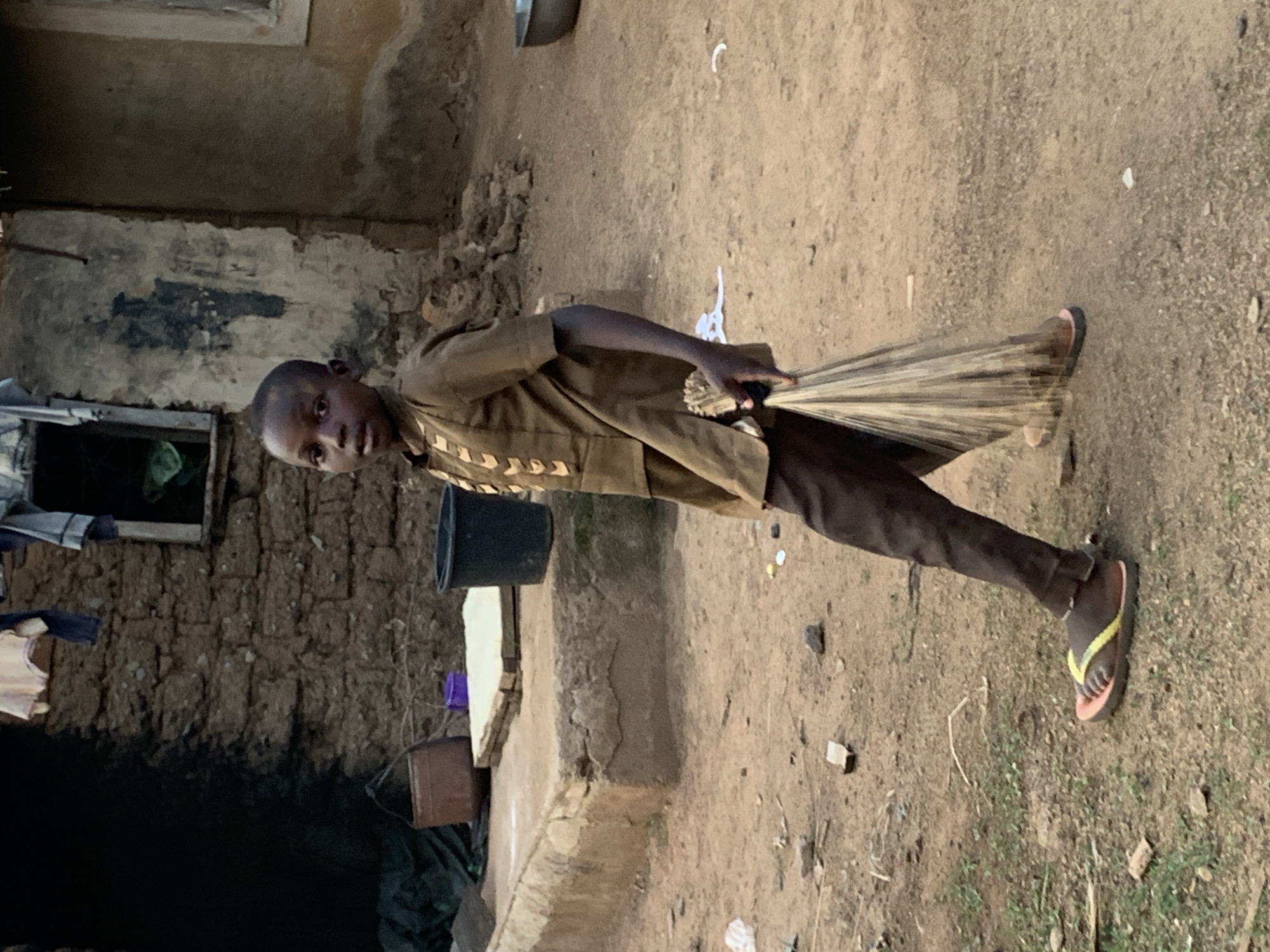 A young boy sweeps the dirt ground in Agban, Nigeria. The boy is wearing a brown shirt and pants and flip-flops. He is holding a straw broom. Behind him is a building made of mud brick. The ground is dirt and debris is scattered across it.