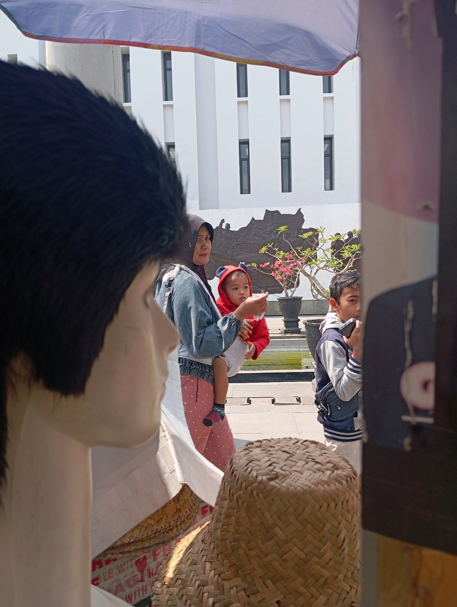 In Blitar, Indonesia, a scene unfolds outdoors, possibly in a public space, in front of a modern white building with repeating rectangular windows. In the foreground, a large black wig and part of a mannequin's face obscure the left side of the image. To the right of the mannequin, a woman wearing a headscarf and denim jacket holds a baby dressed in a red hoodie. The baby appears to be reaching out. Behind them, a young boy in a blue and grey jacket is looking at his phone. In the mid-ground, a small tree with pink flowers is in a pot, and a water feature is visible. A straw hat sits in the foreground, and a large umbrella extends overhead. The overall impression is of a casual outdoor gathering.
