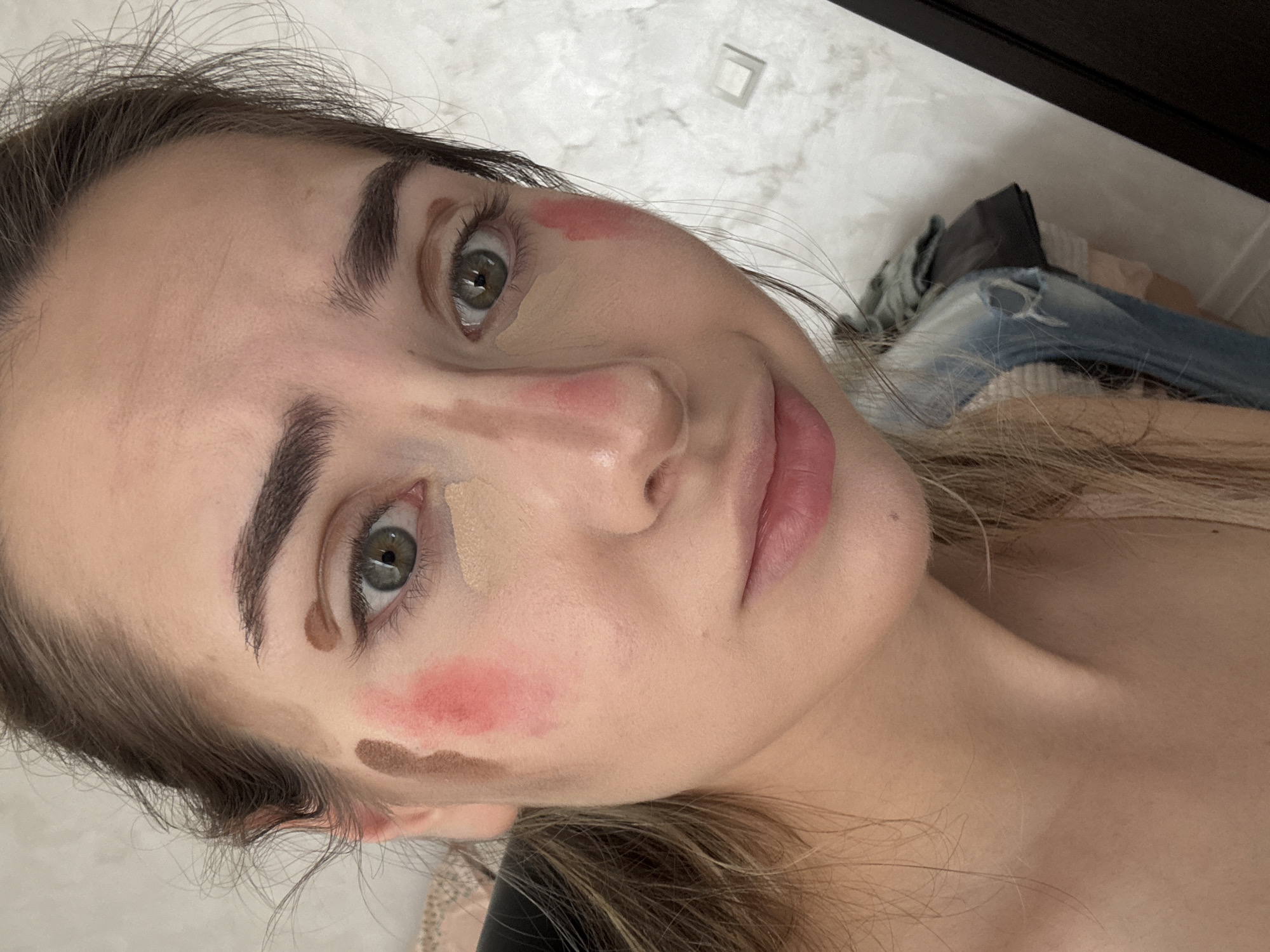 A close-up, slightly angled portrait of a young woman with fair skin and brown hair pulled back from her face. Her face displays makeup application at various stages, including patches of concealer under one eye and on the forehead, pink blush on her cheeks, and brown contouring lines along her nose and jawline. The woman has green eyes and thick, dark eyebrows. The background appears to be a cluttered interior space with a white wall, a dark wooden cabinet, and piles of clothing. Location cannot be determined from the image.