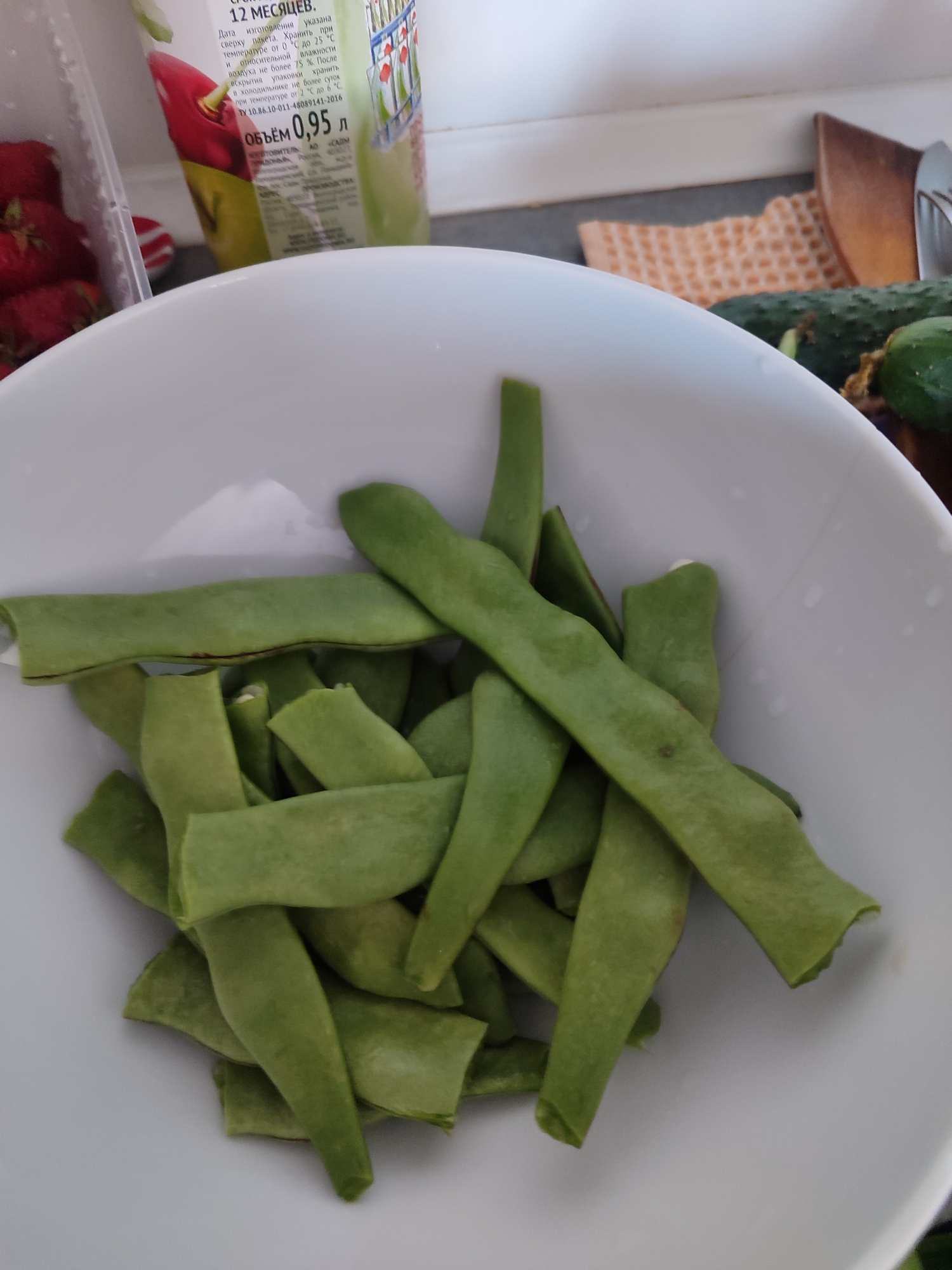 The image shows a white bowl filled with cut green beans. In the background, out of focus, are some strawberries in a clear plastic container, a carton of juice with Cyrillic writing, and some cucumbers. The scene appears to be indoors, possibly a kitchen counter. There are no people or actions visible. The location in Sochi, Russia, is not discernible from the image.