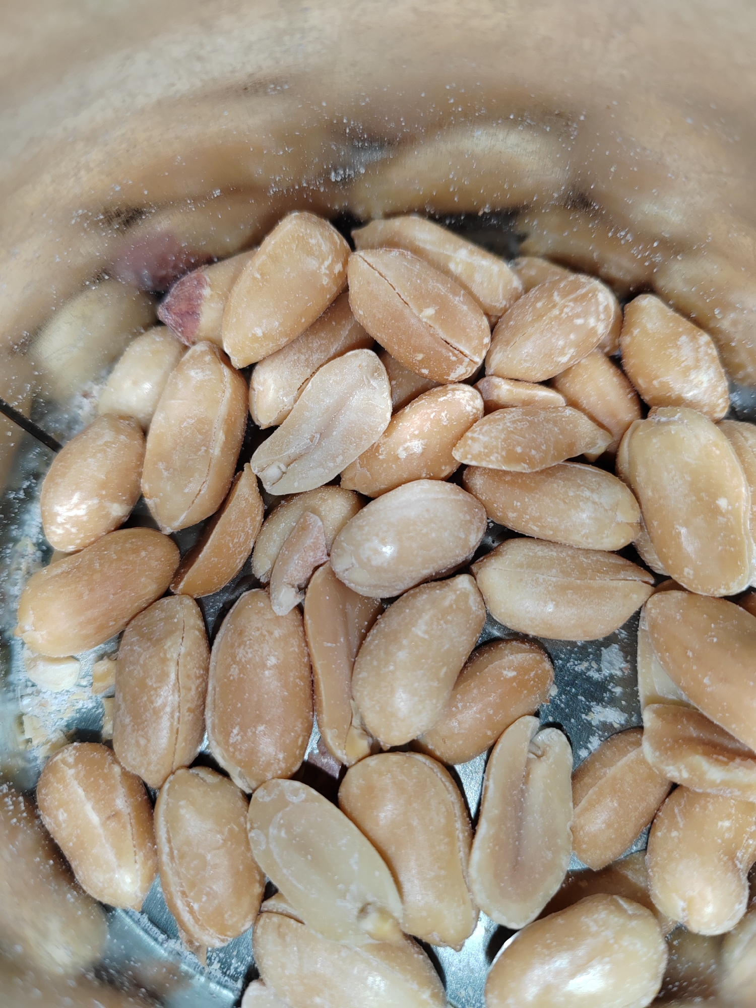 The image is a close-up, top-down view of a pile of shelled peanuts. The peanuts are light brown and appear to be coated in a light dusting of what could be salt or powdered sugar. Some of the peanuts are whole, while others are broken into halves. They are contained within a metallic bowl or container, the interior of which is visible at the bottom of the frame and shows a reflective, textured surface. The lighting is diffused, creating soft shadows and highlighting the texture of the peanuts. The background is blurred and out of focus, suggesting a shallow depth of field. There are no people or other objects visible in the image besides the peanuts and the container. The location is Unknown, Russia.