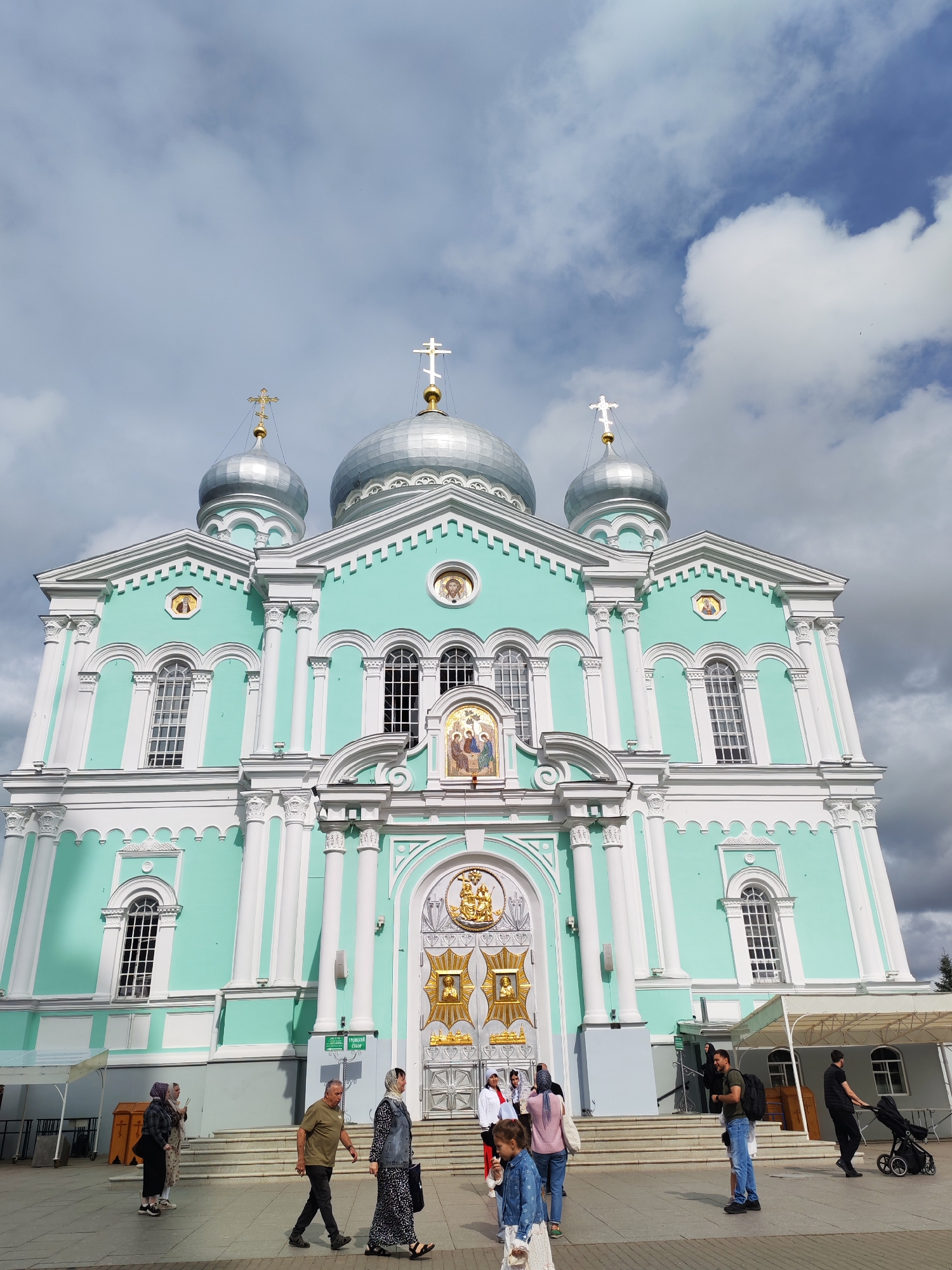 In Diveyevo, Russia, the Transfiguration Cathedral stands majestically under a cloudy sky. The cathedral is a striking edifice painted in a mint green and white color scheme, featuring multiple silver domes topped with gold crosses. Its facade is adorned with ornate columns, arched windows, and golden decorative elements around the main entrance.
In front of the cathedral, a group of people are seen walking. One man, wearing a green shirt and dark pants, walks towards the left of the frame. To his right, a woman in a black and white patterned dress and a denim vest walks past. A young child in a blue dress is also visible in the foreground, facing away from the camera. Other individuals are scattered around, some appearing to be visitors or pilgrims. Several temporary white awnings are set up near the entrance of the cathedral. The ground is paved, and some trash bins are visible near the stairs leading to the cathedral. The overall scene captures a moment of public access to a significant religious site.