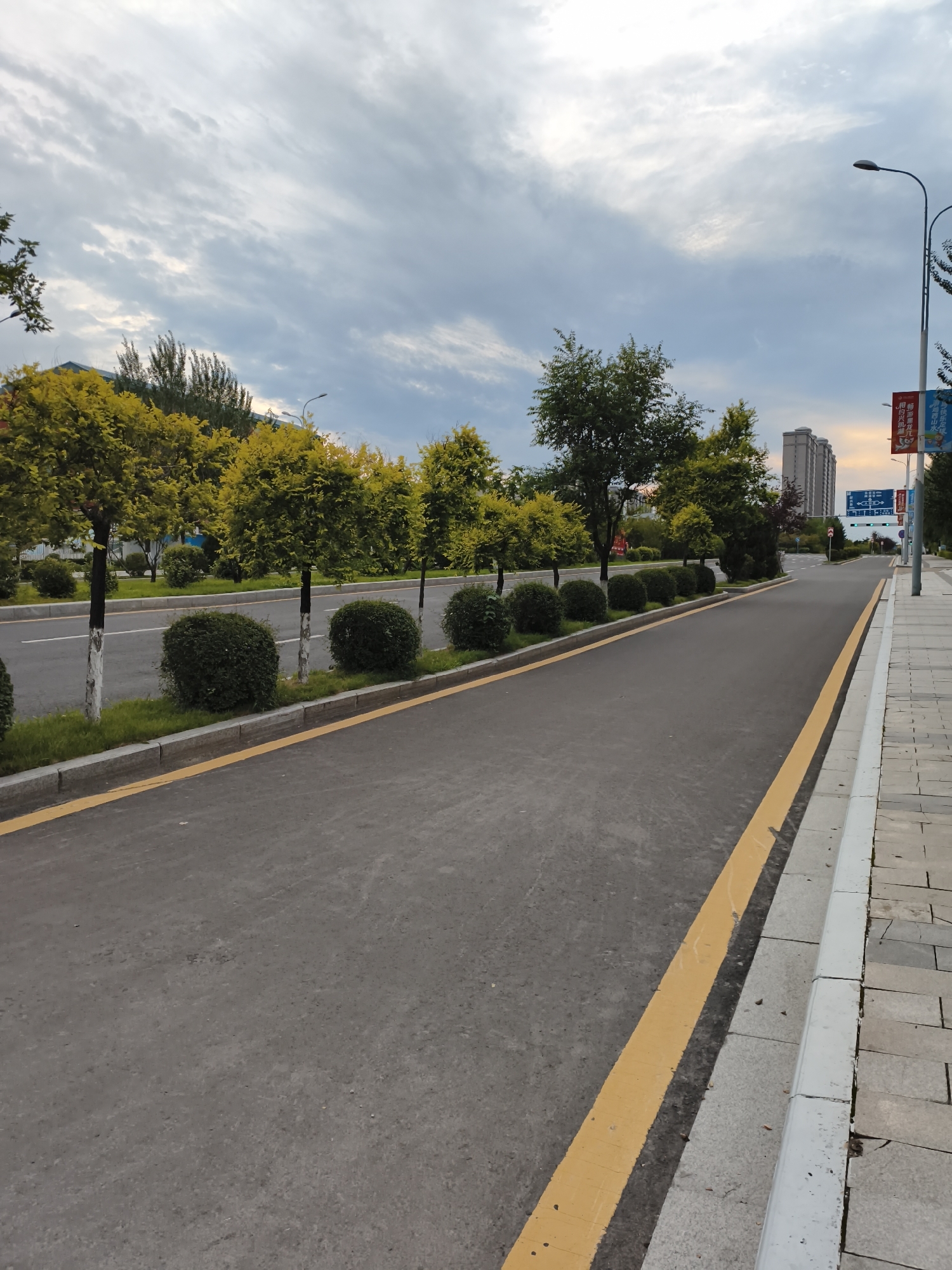 The image shows a street scene in Jiguan District, China. The street is paved with asphalt and has a yellow line marking the edge. On the left side of the street, there is a row of neatly trimmed trees and bushes. The trees are planted in a line along the sidewalk. On the right side of the street, there is a sidewalk made of stone pavers. In the background, there are several buildings, including a tall residential building. The sky is cloudy, and the light suggests it is either early morning or late afternoon. There are streetlights visible along the road, and some banners are hanging from a pole on the right side of the image. No people or vehicles are present in the image. The scene appears to be a well-maintained urban environment.