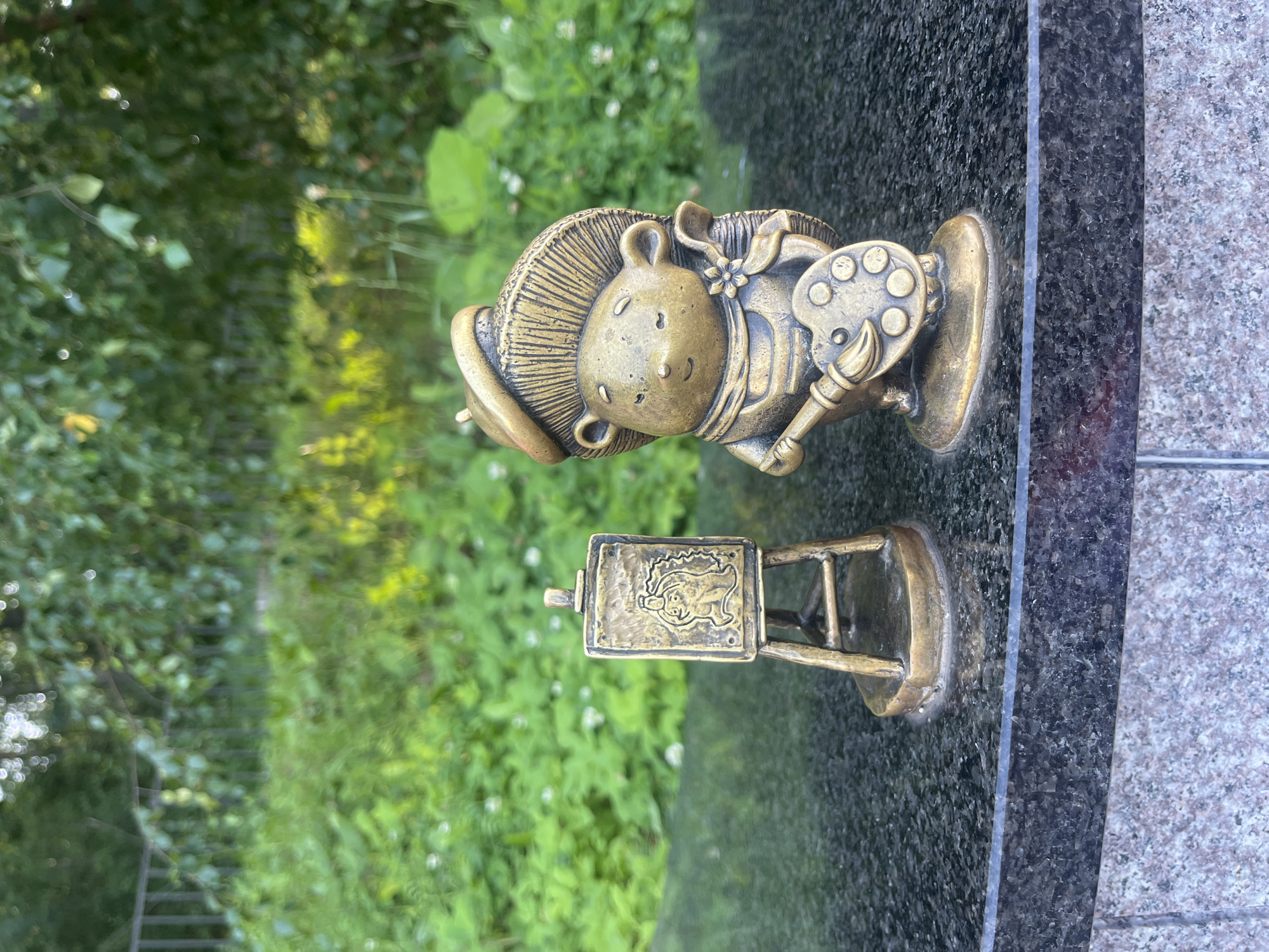 Two bronze figurines are placed on a dark granite surface in front of lush green foliage. To the left, a miniature easel holds a painting of a face. To the right, a hedgehog figurine, dressed in a beret and overalls, holds a paintbrush and a palette. The scene appears to be outdoors, possibly in a park or garden, with soft sunlight filtering through the leaves. The hedgehog figurine is a well-known landmark in Svetlogorsk, Russia, often referred to as the "hedgehog artist."