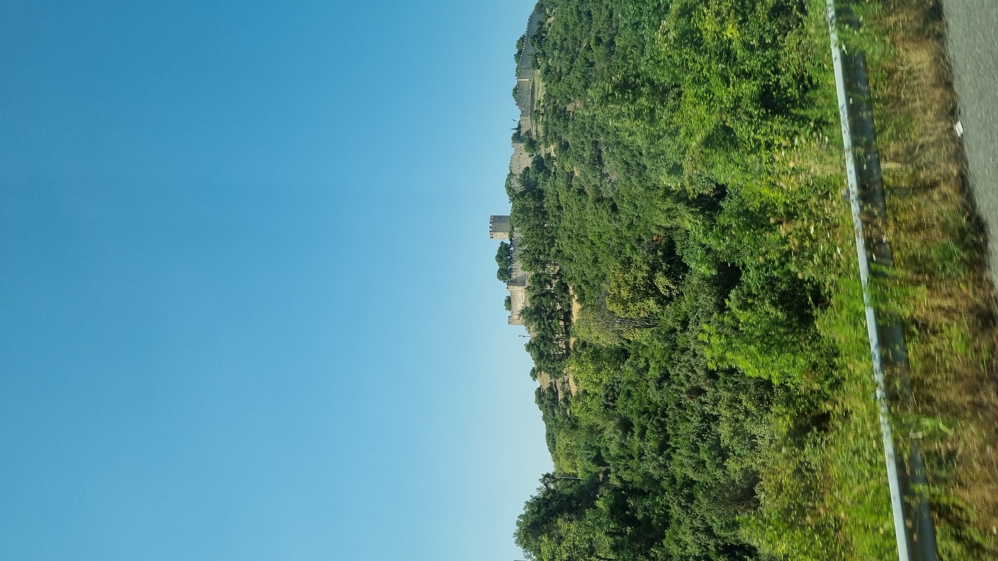A wide-angle shot shows a castle on a hilltop overlooking a lush green landscape under a clear blue sky. The castle, constructed of stone, features crenellated walls and a prominent tower. It is surrounded by trees, with more dense foliage covering the slopes of the hill. In the foreground, a metal guardrail runs horizontally across the bottom of the frame, separating the road from the vegetation. The scene is likely located in or near Neos Panteleimonas, Greece, given its architectural style and the Mediterranean vegetation.