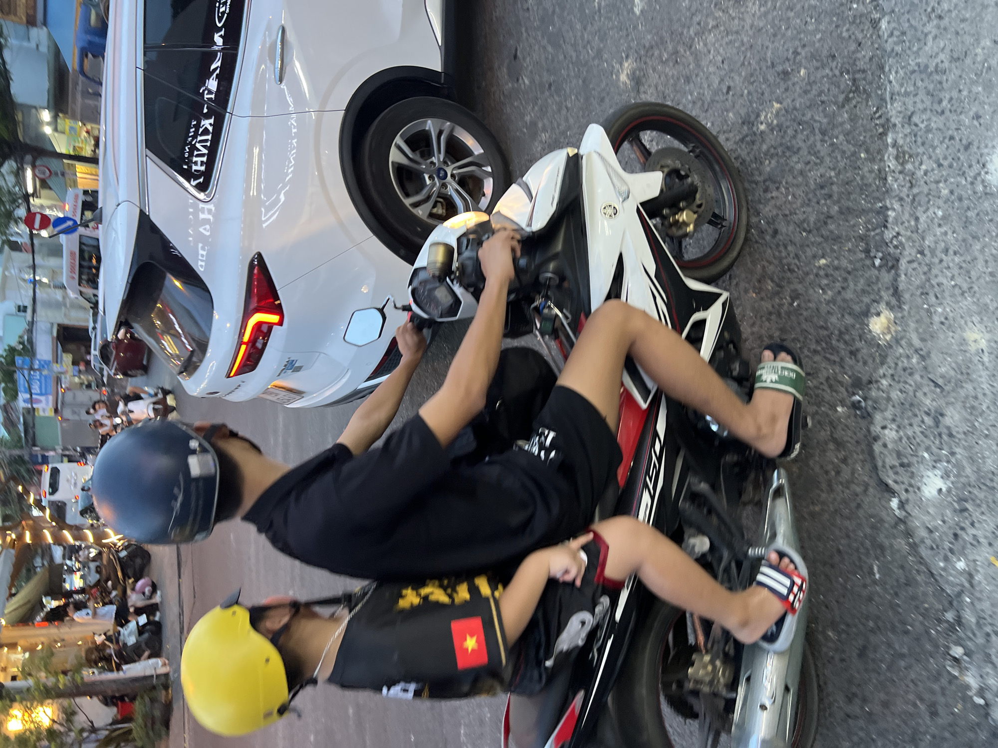Two individuals, likely an adult and a child, are riding a motorcycle on a city street in Nha Trang, Vietnam. The person in front, presumably the adult, is wearing a black t-shirt and a dark blue helmet. The child in the back is wearing a black t-shirt with yellow accents and a red and yellow Vietnamese flag emblem on the left sleeve, and a bright yellow helmet. Both are seated on a white, red, and black motorcycle. The child is wearing red, white, and blue striped sandals, while the adult is wearing green and black slide sandals. They appear to be moving through moderate traffic, as evidenced by a white SUV to their right and other vehicles and pedestrians in the background. The street is paved, and there are buildings and streetlights visible along the sides of the road, suggesting an urban environment. The lighting indicates it is either late afternoon or early evening.