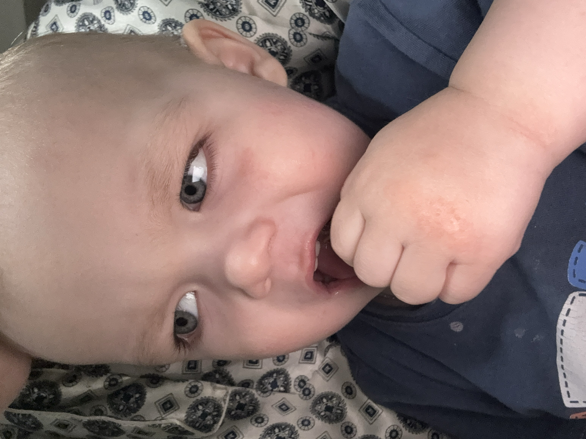 A baby lies on patterned fabric wearing a blue shirt and putting a hand in their mouth. The location is Unknown, Unknown.