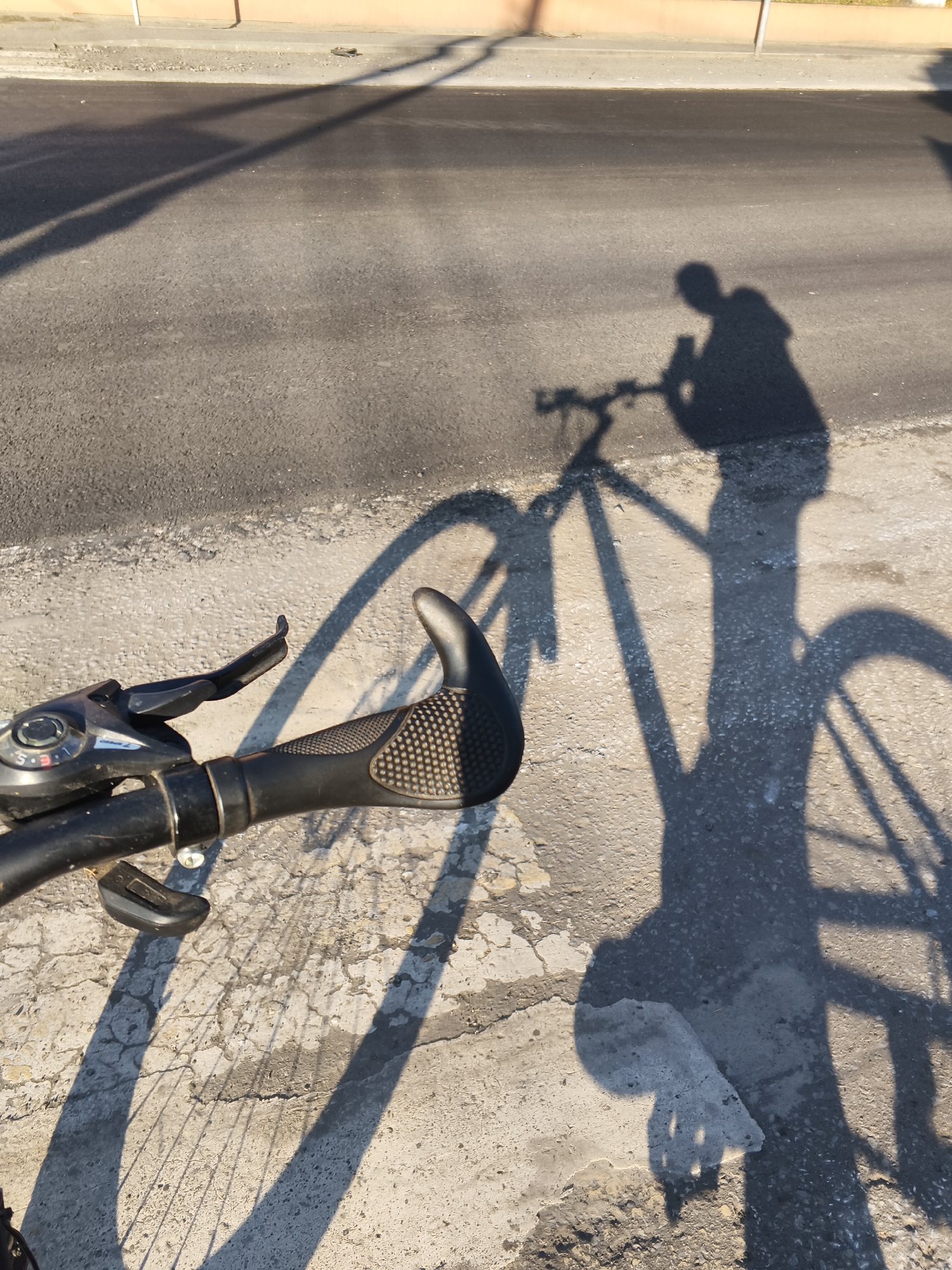 The image shows a close-up view of a bicycle's handlebar and brake lever, with the shadow of the cyclist and bicycle cast on asphalt and concrete pavement. The scene is outdoors, likely on a street or path in the city of Mokshan, Russia, given the provided context. The shadow indicates a person is riding the bicycle, as the shape of a human figure and the bicycle's frame, wheels, and handlebars are visible in silhouette. The pavement appears to be a mix of dark asphalt and lighter, cracked concrete. The lighting suggests it is either early morning or late afternoon due to the long shadows.