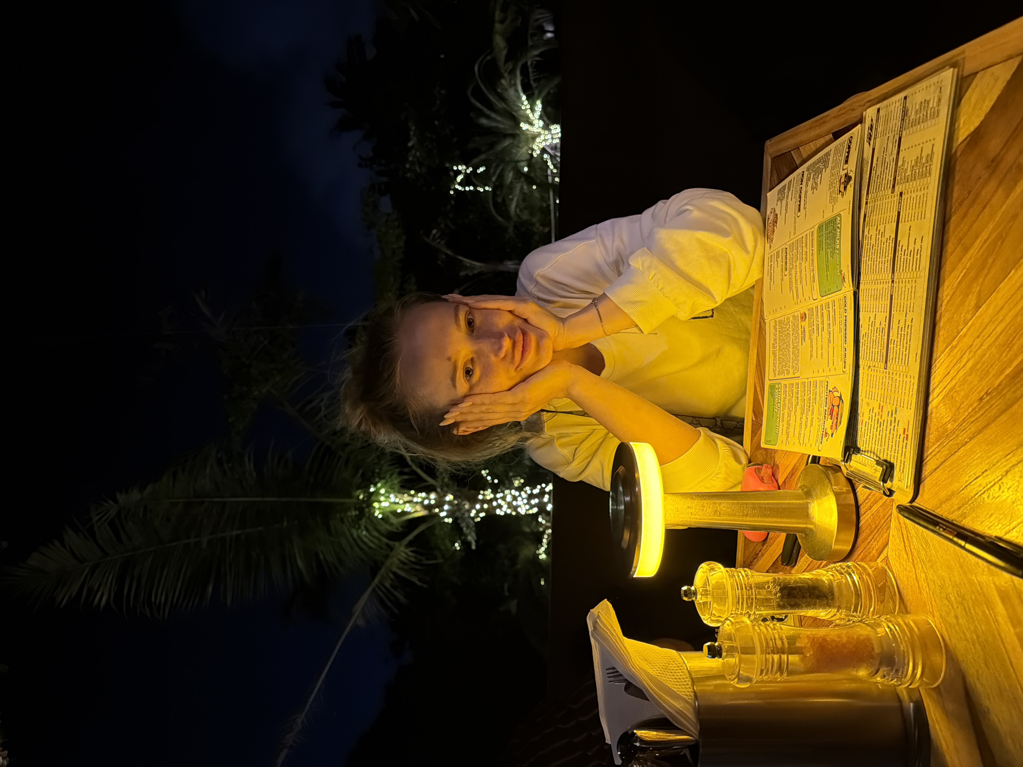A young woman with blonde hair pulled back sits at a wooden table outdoors at night. She is wearing a white long-sleeved shirt and has her hands on her cheeks, smiling at the camera. In front of her is a lit table lamp, salt and pepper shakers, a cutlery holder, and menus. In the background, palm trees decorated with white fairy lights are visible against a dark sky. The scene appears to be in Mengwi, Indonesia.
