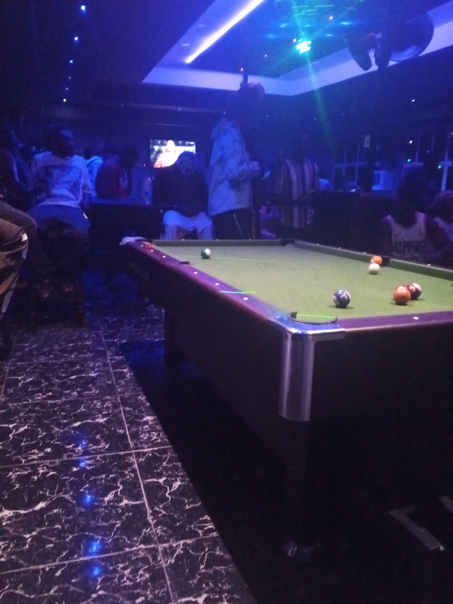 **Location:** A dimly lit bar/entertainment establishment in Lokoja, Nigeria.
**People:** Several individuals are present, mostly in the background. Some are sitting, others are standing.
**Subjects:** A billiard table dominates the foreground.
**Objects:** Billiard balls scattered on the table. The floor is tiled with a black and white pattern. Overhead lighting. A screen displaying something is visible in the background.
**Actions:** People are socializing and observing the billiard game.
**Interactions:** People are gathered around the billiard table.
**Environment:** The scene is dimly lit with blue and white lights, creating a nightlife atmosphere.