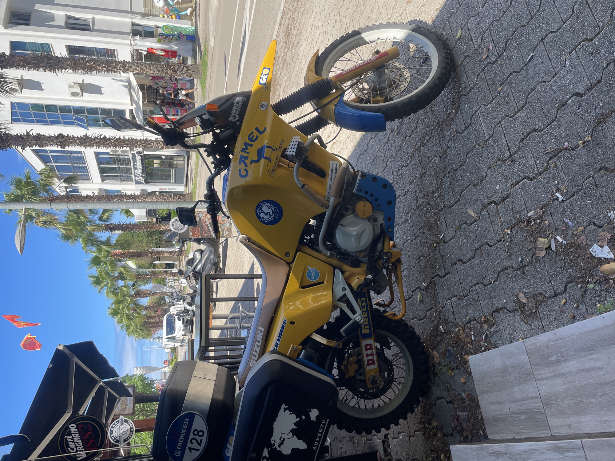 An overcast, sunny day in Bar, Montenegro, features a bright yellow Suzuki motorcycle parked on a paved walkway next to a cafe. The motorcycle, adorned with various decals including "Camel," "GEO," "NASA," and a world map on its saddlebag, is the primary subject. A black top case is mounted on the rear. In the background, palm trees line the street, and a white building with multiple windows stands prominently. Several parked scooters and a white van are also visible. Red flags flutter in the sky above a black awning that covers the cafe. The walkway is strewn with fallen leaves and debris.