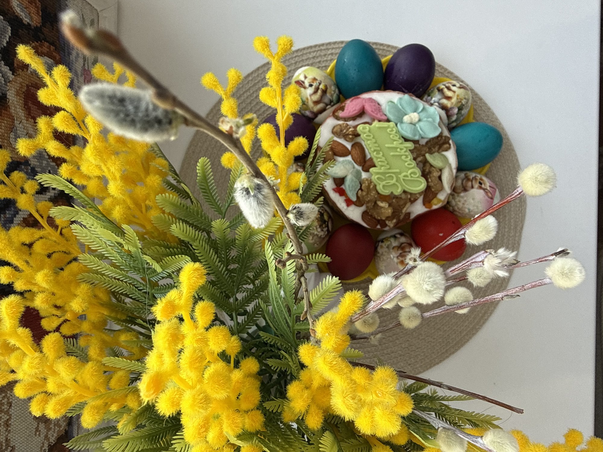 The image presents a close-up shot of a floral arrangement, presumably for Easter celebration, against a neutral background. The arrangement is dominated by vibrant yellow mimosa flowers and pussy willow branches, creating a textured foreground. The centerpiece of the arrangement is an Easter cake (kulich) decorated with almonds, nuts, and confectionery, and topped with a green label written in Cyrillic script, indicative of Russian or Slavic origin. Surrounding the cake are dyed Easter eggs in various colors, including blue, purple, and red. Some eggs have decorative patterns on them. The arrangement sits on a round, woven mat. The presence of the Easter cake and eggs suggests a holiday celebration. The Cyrillic inscription on the cake's label points to a Slavic cultural context. The absence of people and specific landmarks makes it difficult to pinpoint the location precisely, however, the mention of Bor, Russia suggests that the picture was taken either at this location or it involves a person from the location.