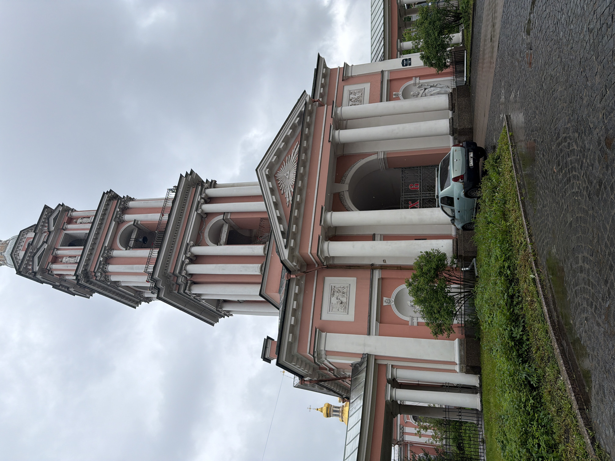 The image shows the Church of St. John the Baptist at Chesme Palace in Saint Petersburg, Russia. The church has a distinctive pink and white facade with classical columns and ornate detailing. A tall, tiered bell tower rises on the left side of the frame. The sky is overcast. A light blue car is parked near the church entrance. A green hedge runs along the side of the building, separating it from a paved area.
