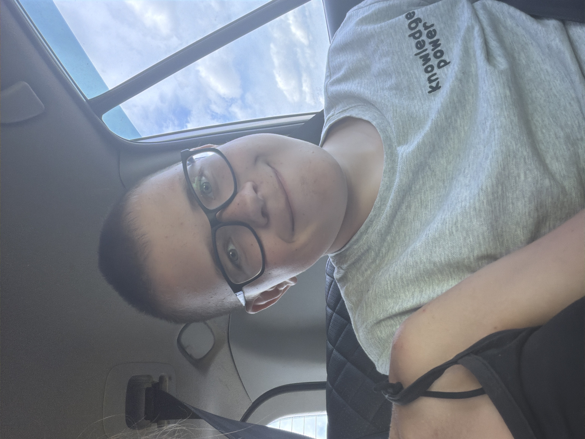 A young person with short hair and glasses is seated in the back of a car, smiling at the camera. The person is wearing a grey t-shirt with the words "knowledge power" printed on it in black. The car's interior is visible, including the patterned seat and a seatbelt. Outside the car window, a cloudy sky is visible. A portion of another person's arm and shoulder wearing a black tank top is in the foreground. The scene appears to be a casual daytime snapshot, likely taken in or near Levyi Salavan, Russia, given the context.