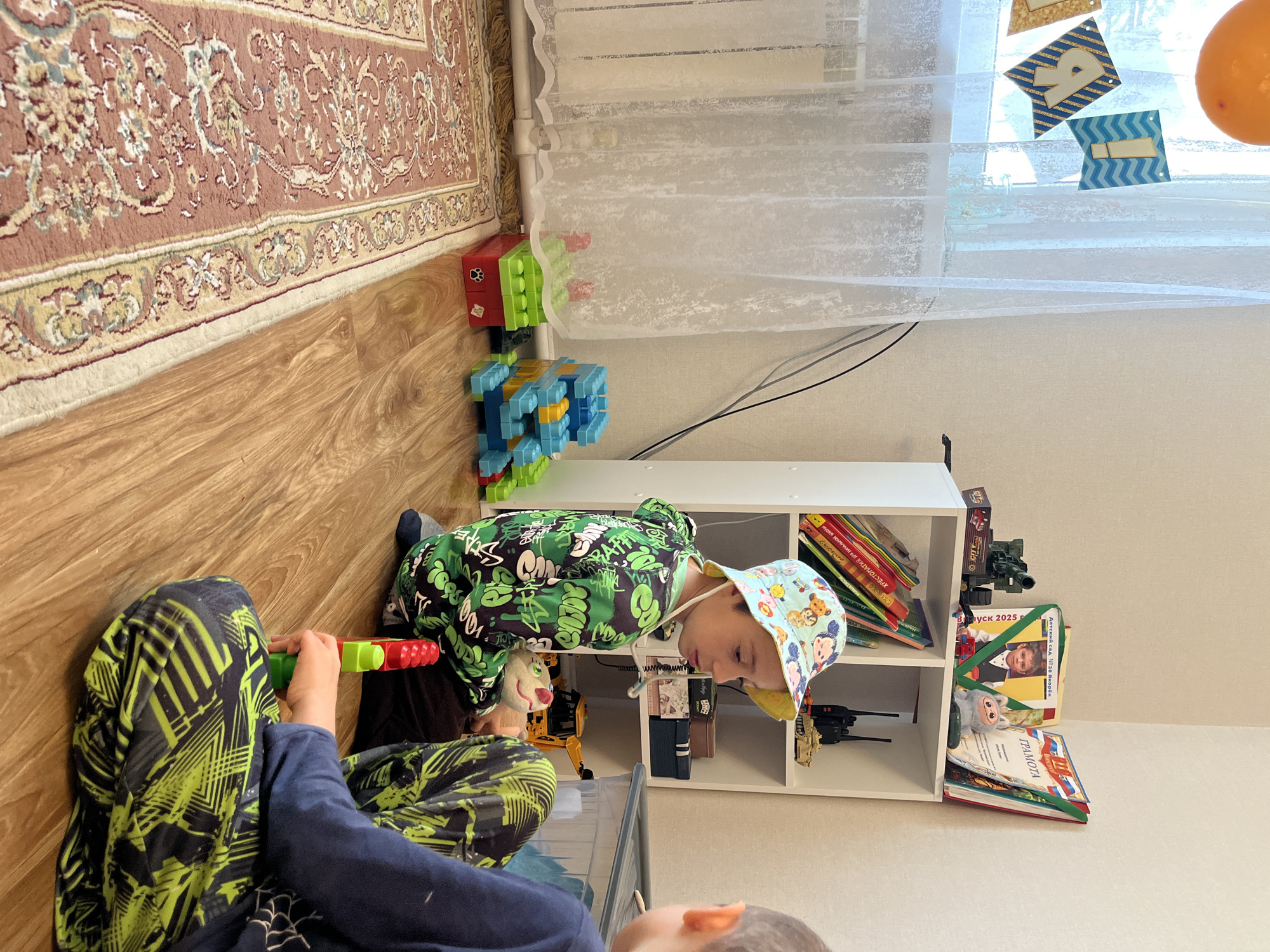 The image depicts an indoor scene with two children present. One child, wearing a green and black hooded jacket and a patterned hat, is holding a toy gun. The other child is partially visible in the foreground. They are sitting on a wooden floor next to a rug with an intricate pattern. A white shelving unit is positioned against the wall, containing books and toys. In the background, there is a window with a sheer white curtain adorned with birthday decorations. The location is Kaspiysk, Russia, based on a book visible on the shelf.