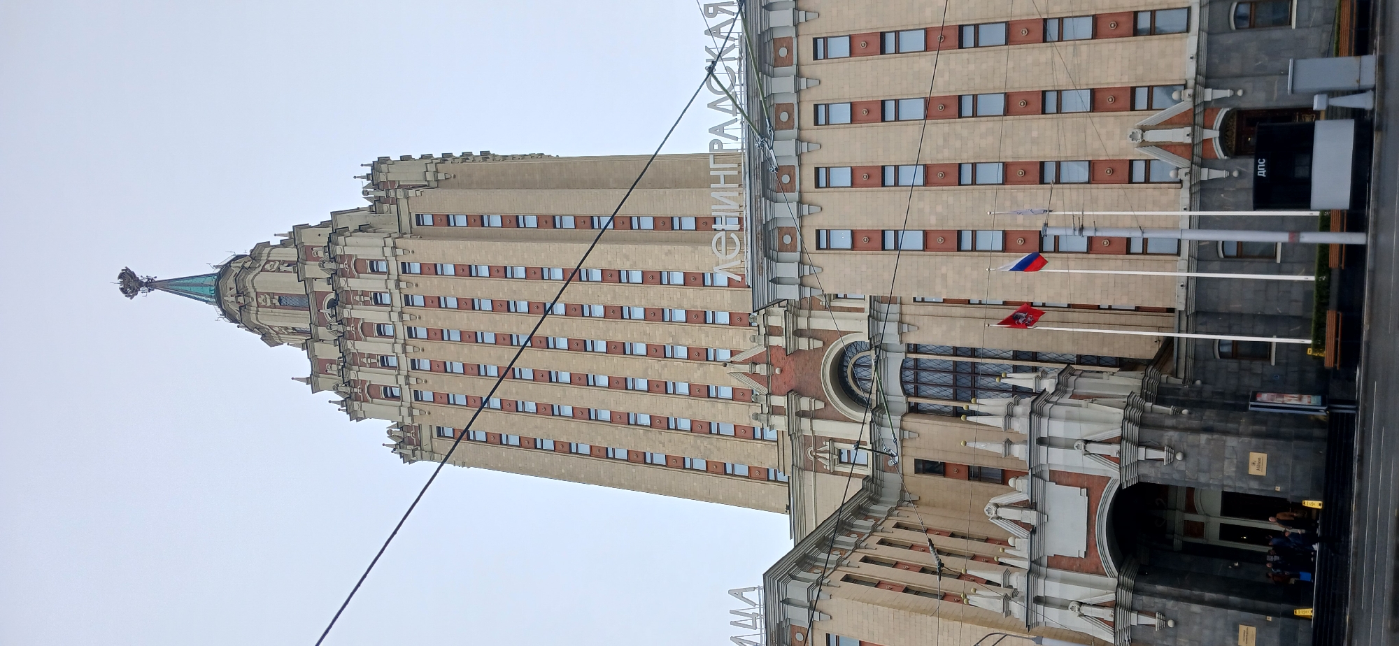 The image shows a tall building with a beige facade and numerous windows arranged in vertical rows. The windows are rectangular and dark, contrasting with the light-colored building material. At the top of the building is a spire-like structure with a green roof and a decorative finial. The building is adorned with architectural details such as cornices, ledges, and decorative elements. On the lower right side of the building, there are flags, including the Russian flag, displayed on flagpoles. A sign with Cyrillic lettering is visible on the side of the building. The building has a prominent entrance with arched windows and decorative stonework. The scene appears to be in an urban setting, possibly a city in Russia. The location is Moscow, Russia.