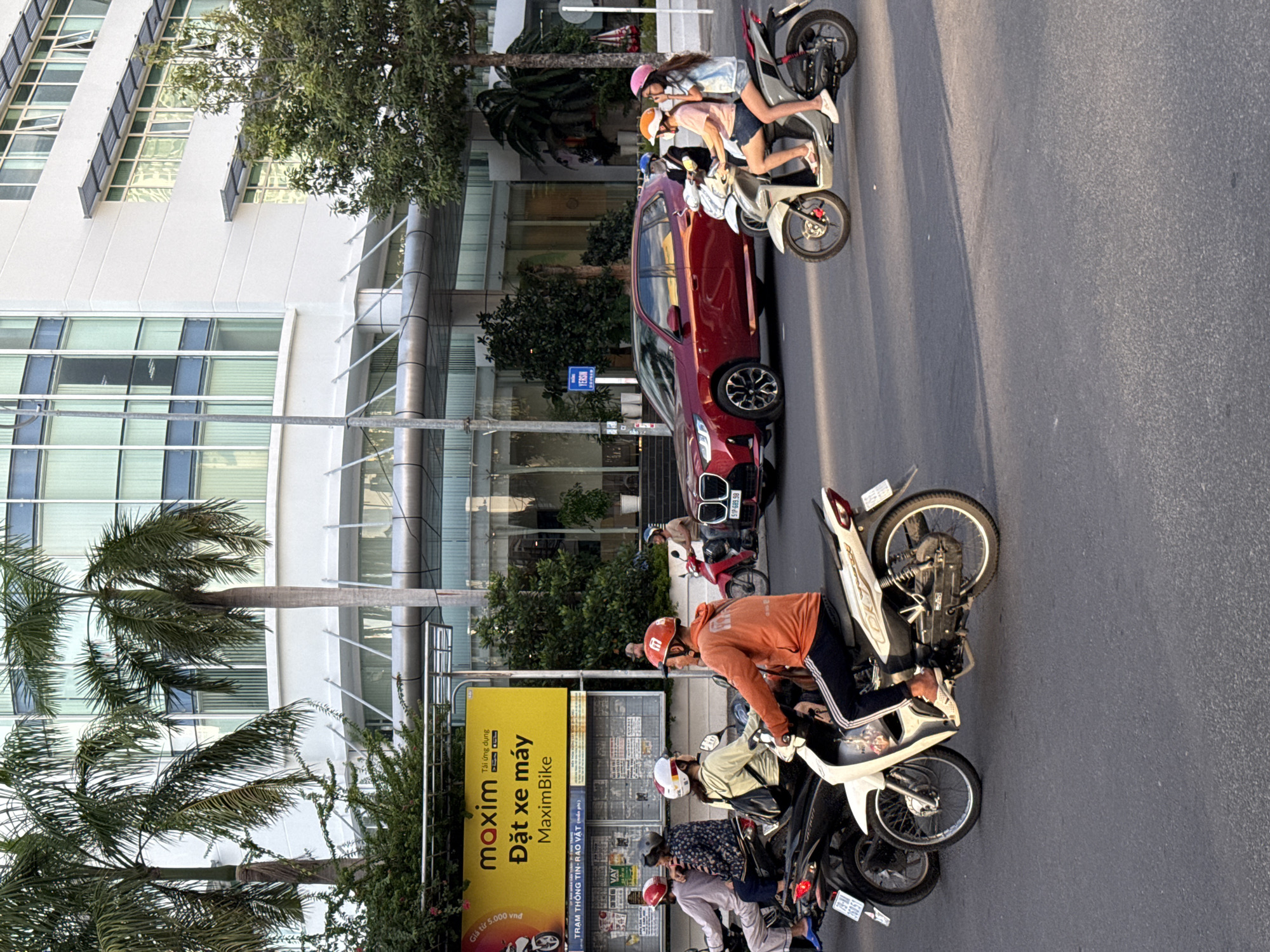 In Nha Trang, Vietnam, on a paved city street, a red BMW SUV drives past a row of motorbikes. Three people ride on two separate motorbikes in the foreground, while two additional people ride on a third motorbike behind them. The riders are all wearing helmets and casual clothing. A bright yellow billboard with Vietnamese text and the Maxim logo is visible in the background. The scene takes place in front of a modern white building with large glass windows, surrounded by palm trees and greenery. The sunlight casts shadows on the street.